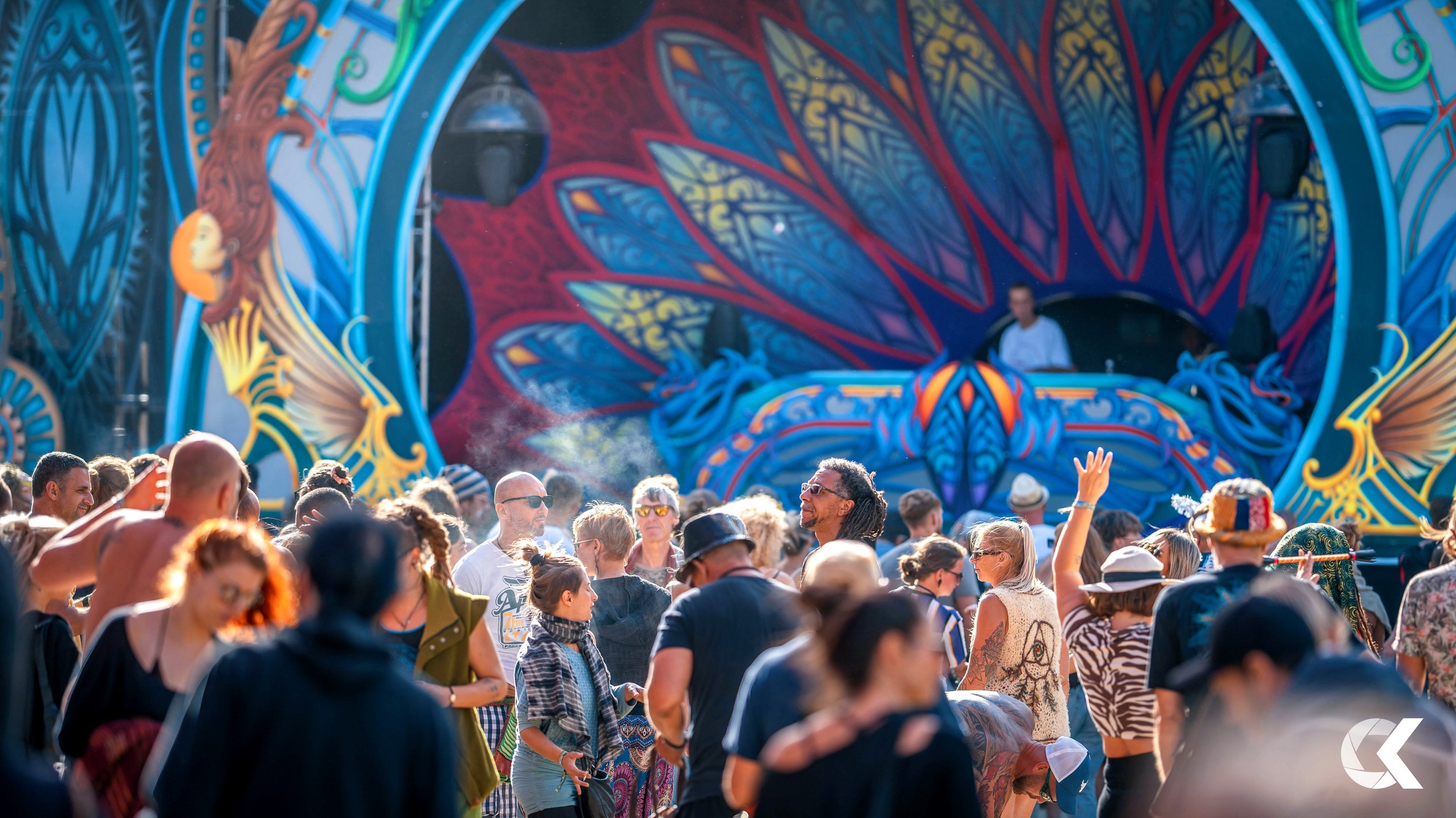 A crowd of people dancing and enjoying a music festival outdoors with a colorful, abstract stage design in the background featuring vibrant patterns and wing motifs.
