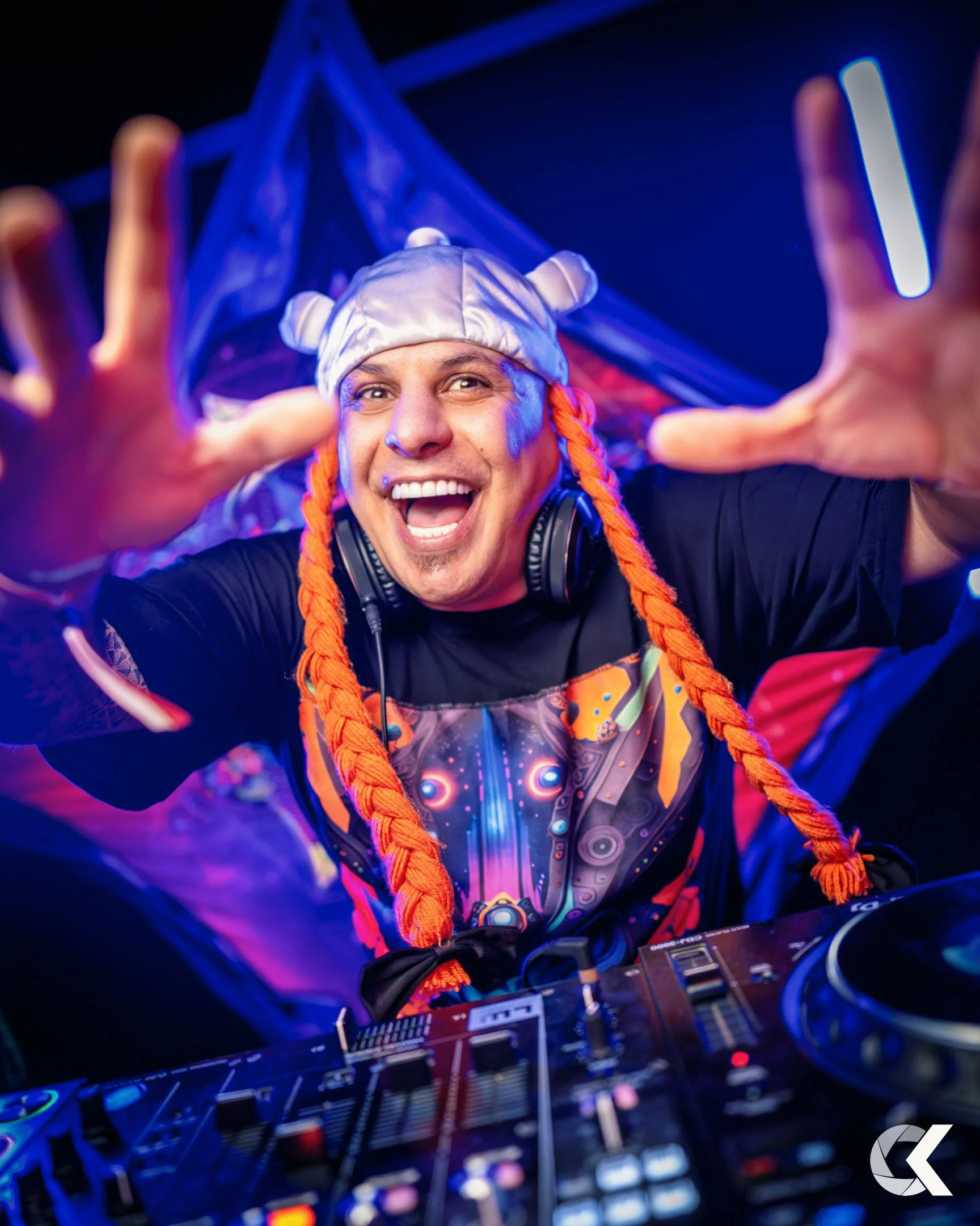 A DJ at a nightclub wearing a silver hat with ears, orange braids, and headphones, smiling and reaching toward the camera with a DJ mixer in front of him, colorful lighting in the background.