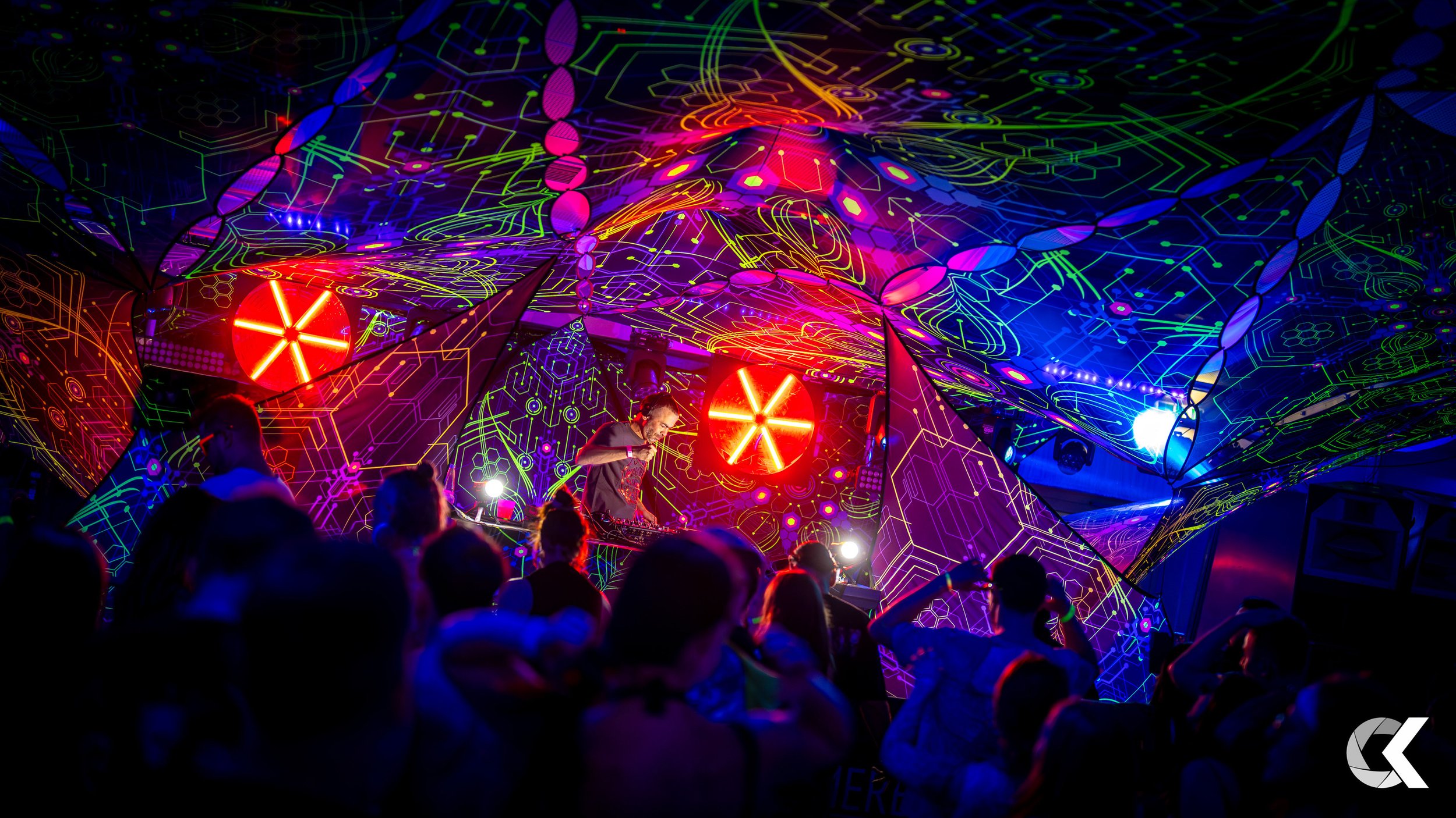 A nightclub or concert scene with a DJ performing on stage, surrounded by vibrant, neon-colored geometric and abstract light patterns on the ceiling and walls. People are dancing and enjoying the music.
