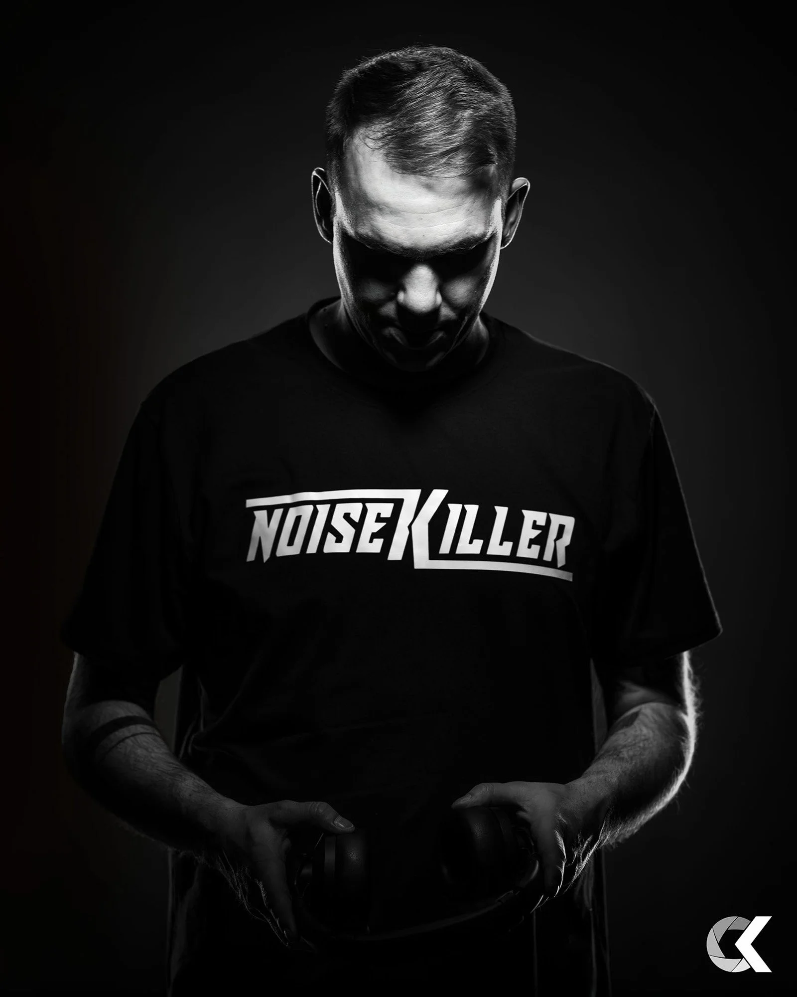 Schwarz-weiß-Foto eines Mannes mit kurzen Haaren, der nach unten blickt und eine schwarze T-Shirt trägt mit dem weißen Text "NOISE KILLER".
