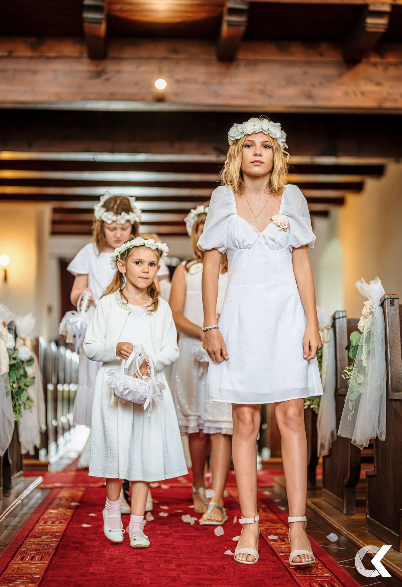 A group of women and girls dressed in white, walking down a red carpet aisle decorated with flowers, during a church ceremony. The women wear white dresses with floral headbands, and the girls hold small pillow-shaped items.