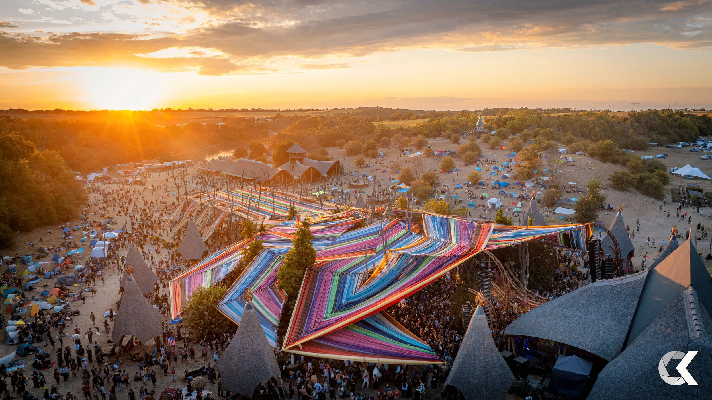 Music festival at sunset with a large crowd, vibrant colorful fabric decorations, and multiple tents and structures in a countryside setting.