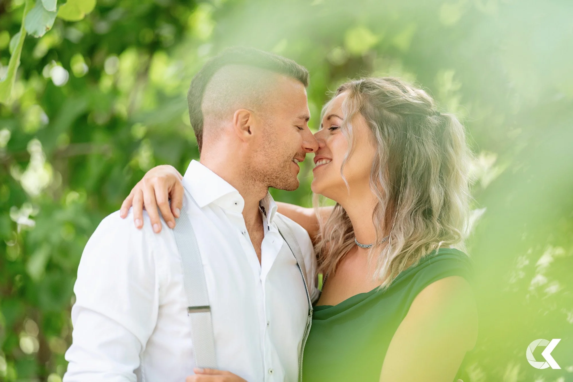 A couple sharing a close embrace outdoors, smiling, surrounded by green foliage.