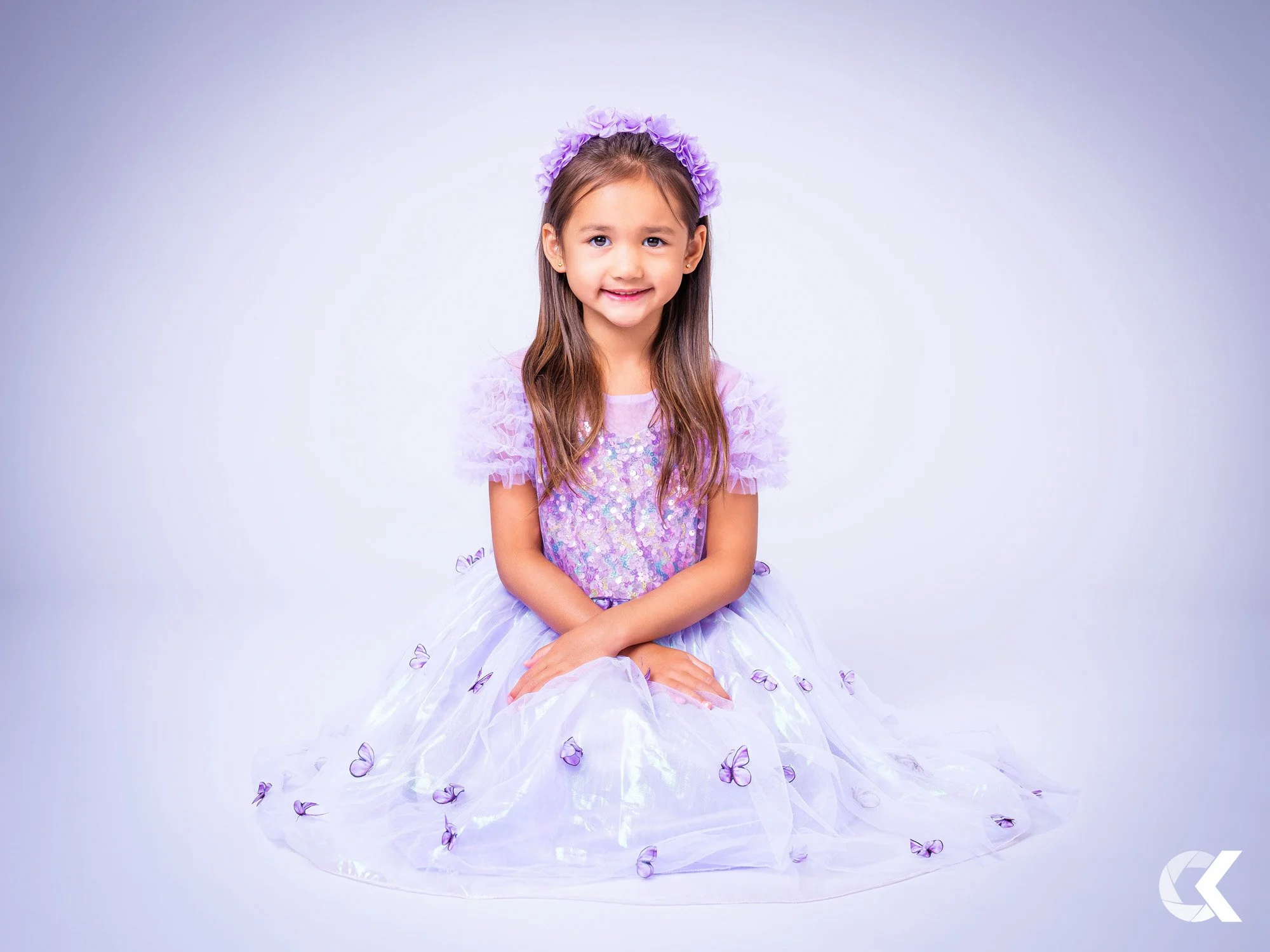 A young girl with long brown hair smiling, dressed in a purple and lavender fairy-like dress with butterfly details, sitting cross-legged on the ground against a plain backdrop.