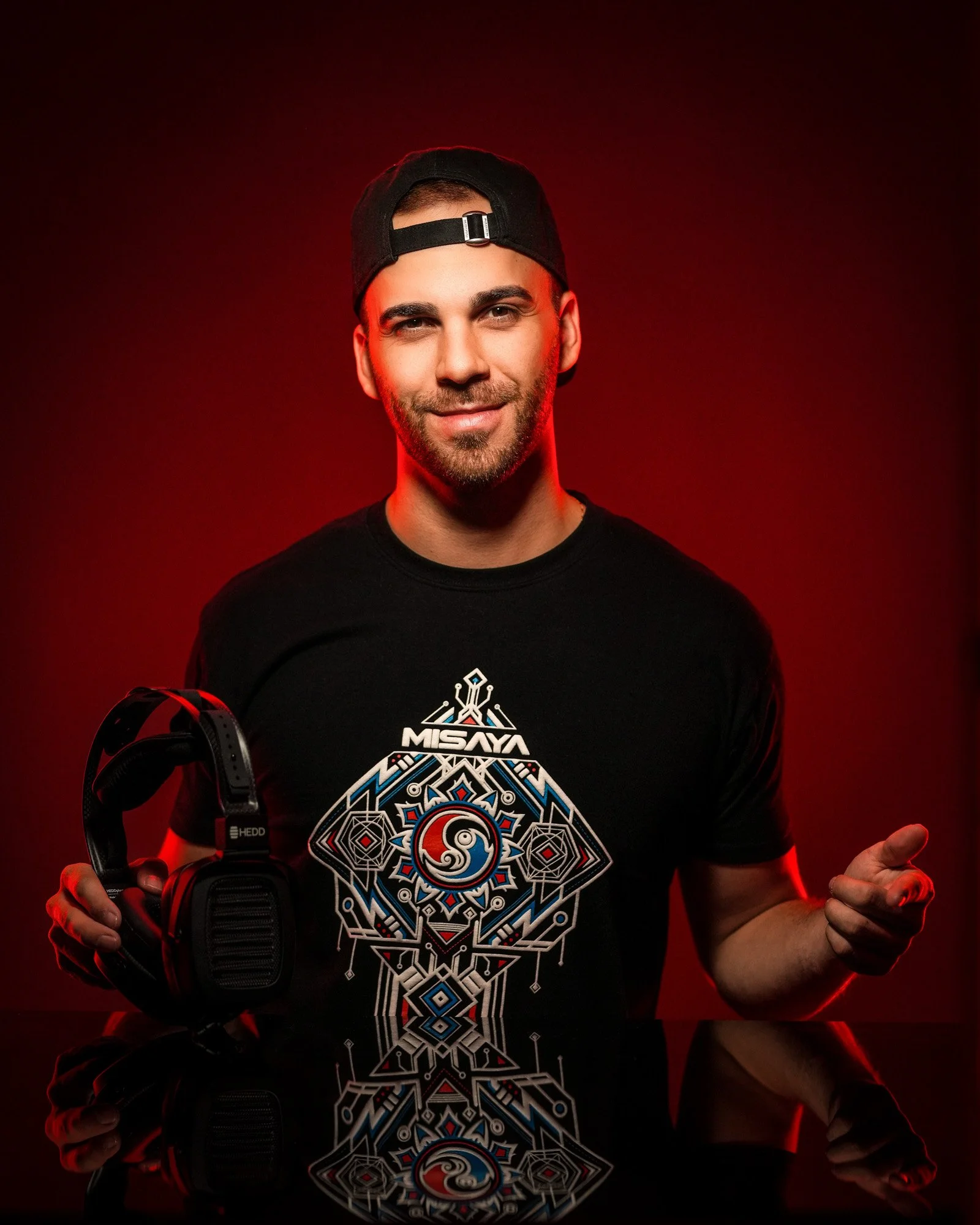 A young man smiling, wearing a black baseball cap backwards and a black t-shirt with a colorful geometric design and the word "MISAYA" on it, holding a pair of black headphones in his left hand against a red background.