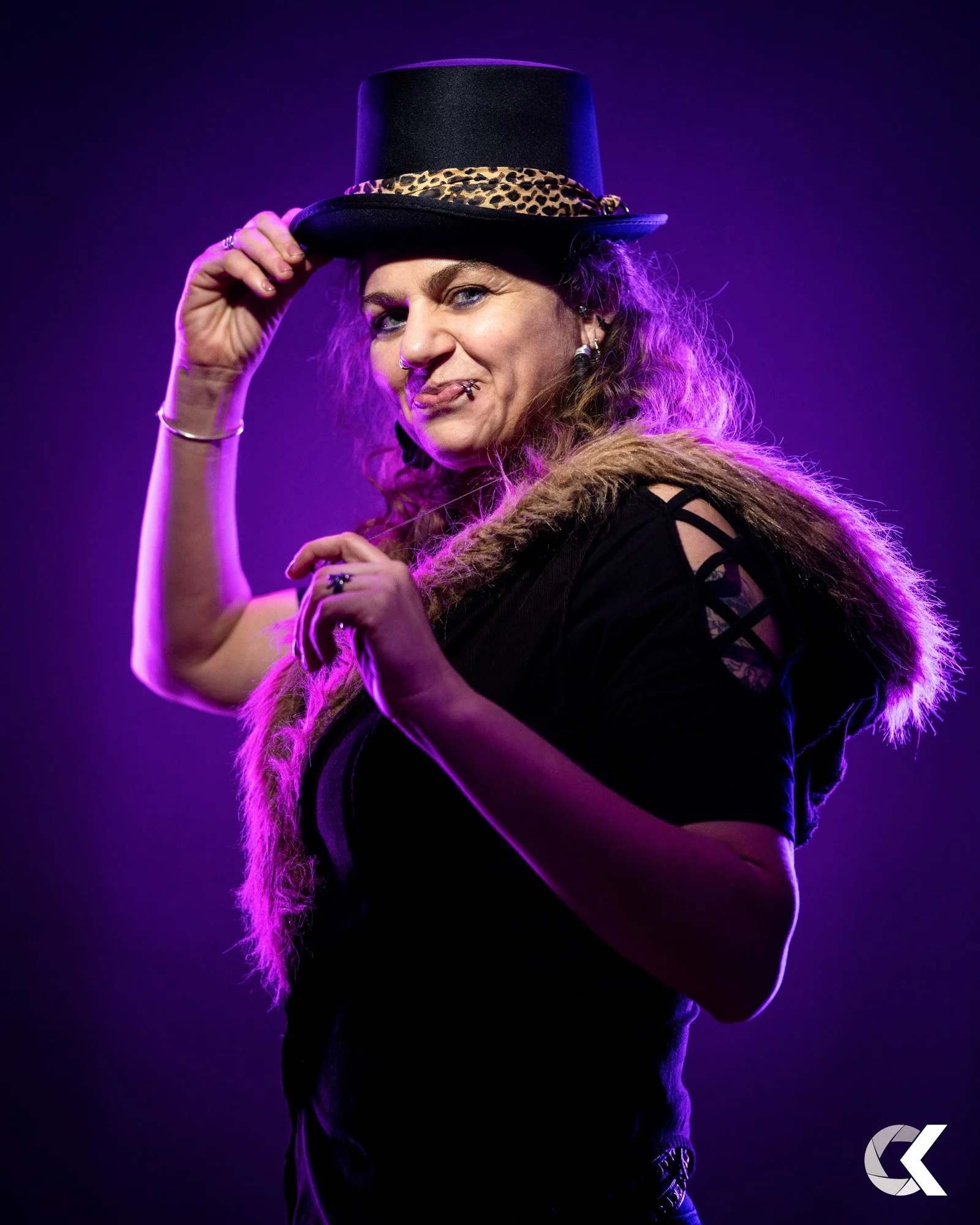 A woman with curly hair wearing a black top with cutout sleeves, a fur stole, and a top hat with a leopard print band, posing against a purple background.