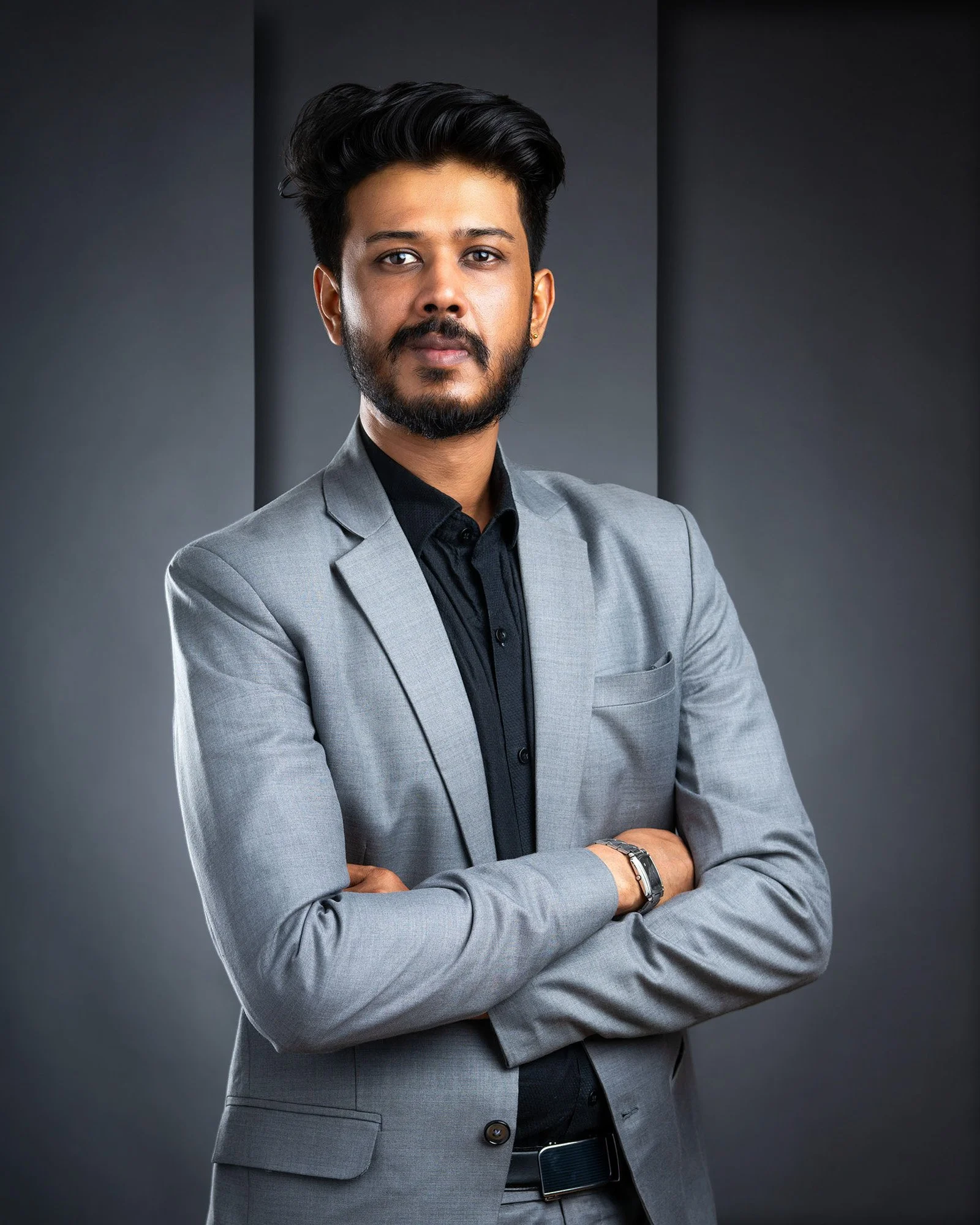 Porträt eines Mannes in Graublazer mit schwarzen Hemd, Arme verschränkt, vor dunklem Hintergrund.