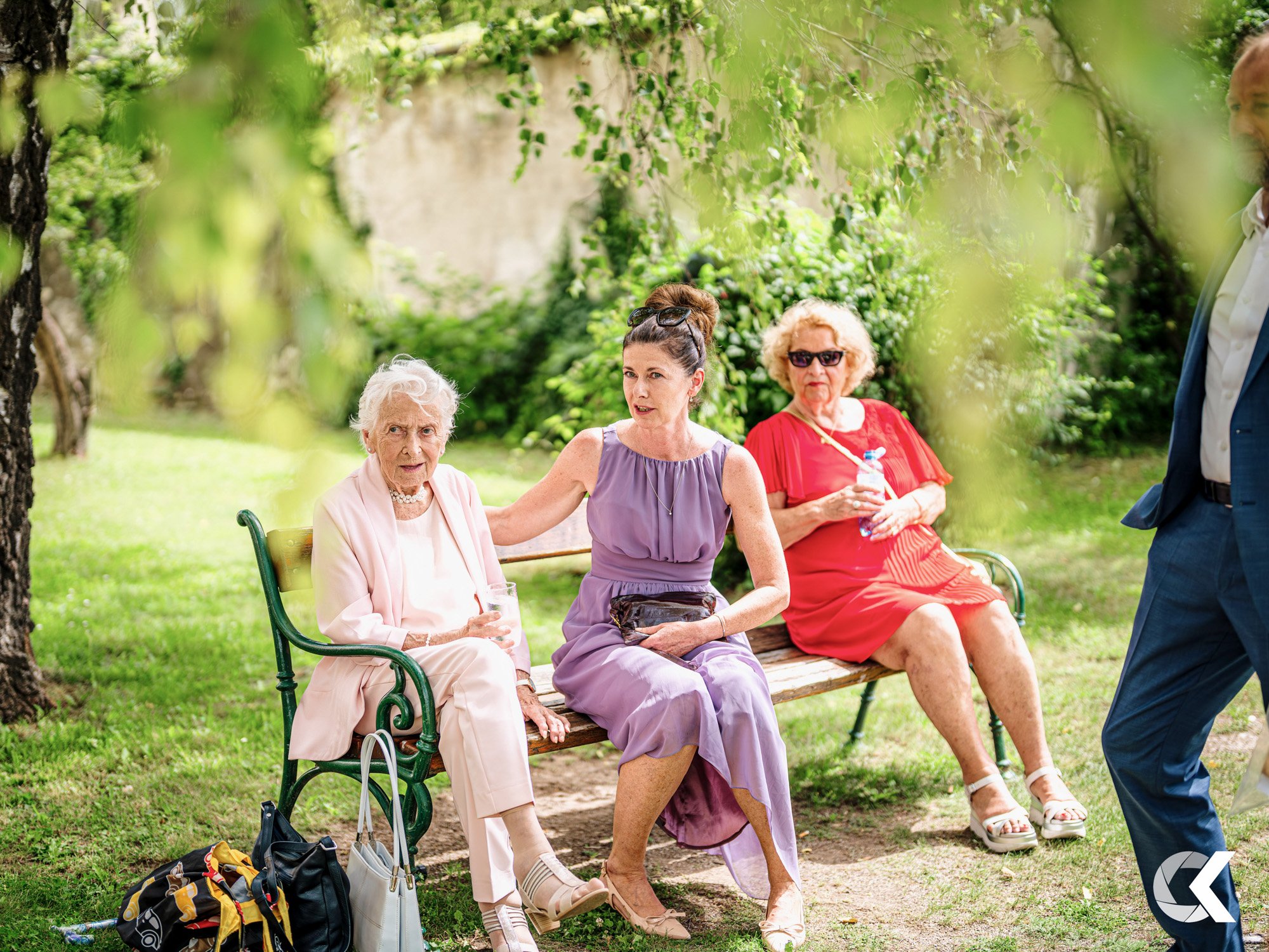 Three elderly women and one younger woman sitting on a park bench surrounded by green trees and grass. The elderly women are holding drinks, and the younger woman has her arm around one of them.