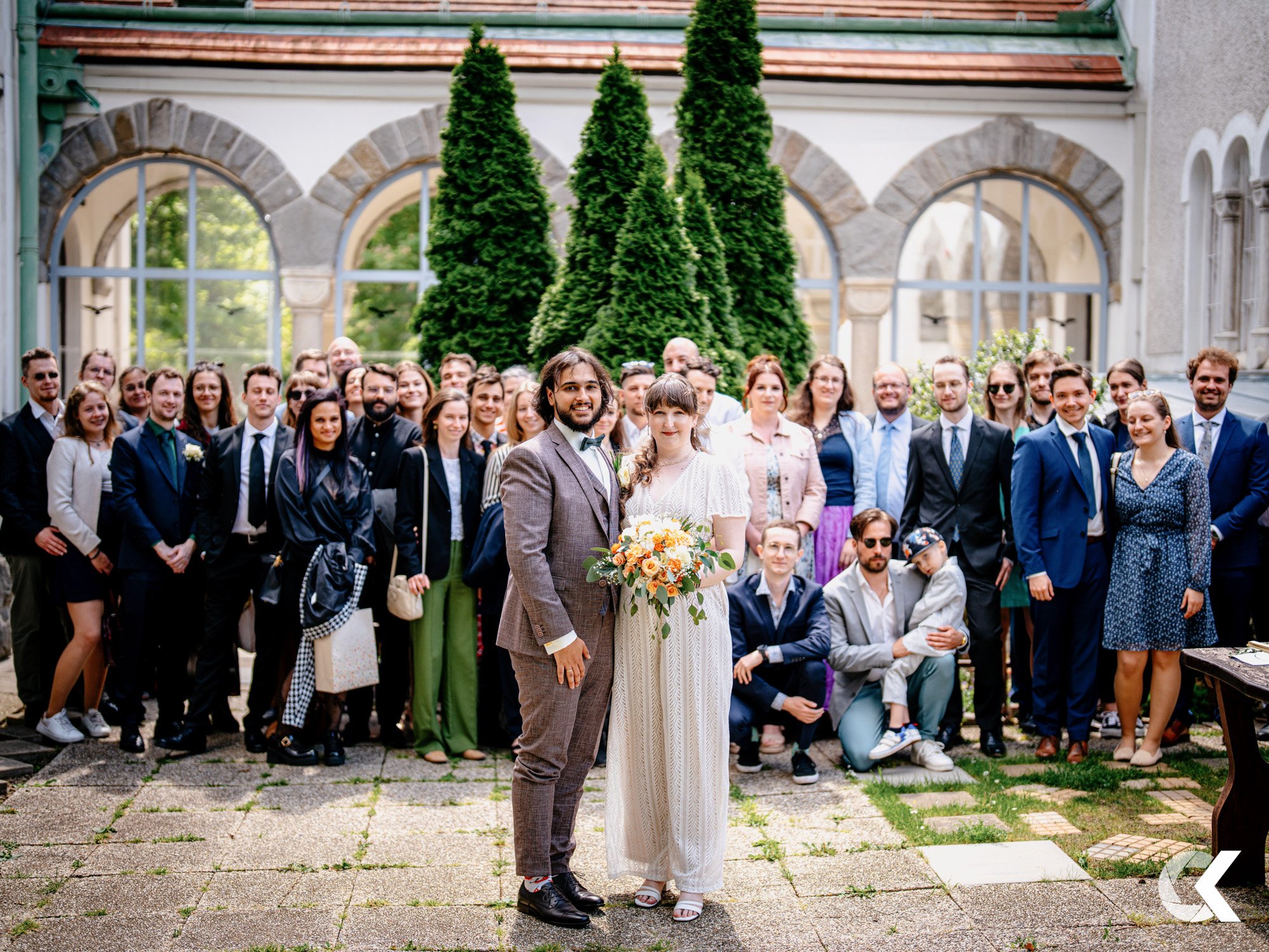 A group of wedding guests gathered outdoors with a bride and groom in front, greenery and arched windows in the background.
