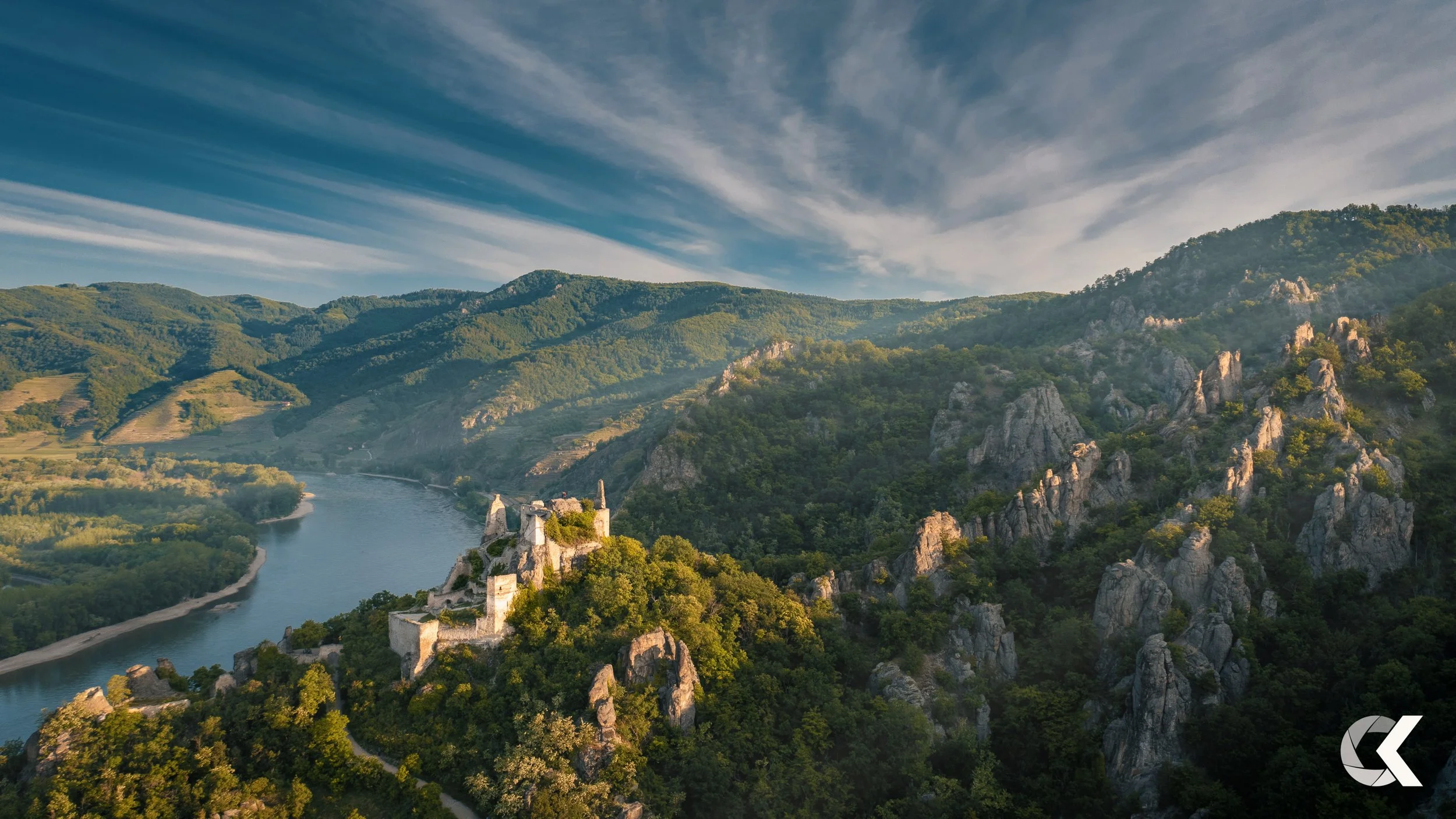 Scenic landscape of a river winding through lush green hills and mountains under a partly cloudy sky, with ruins of an old castle on a hill in the foreground.