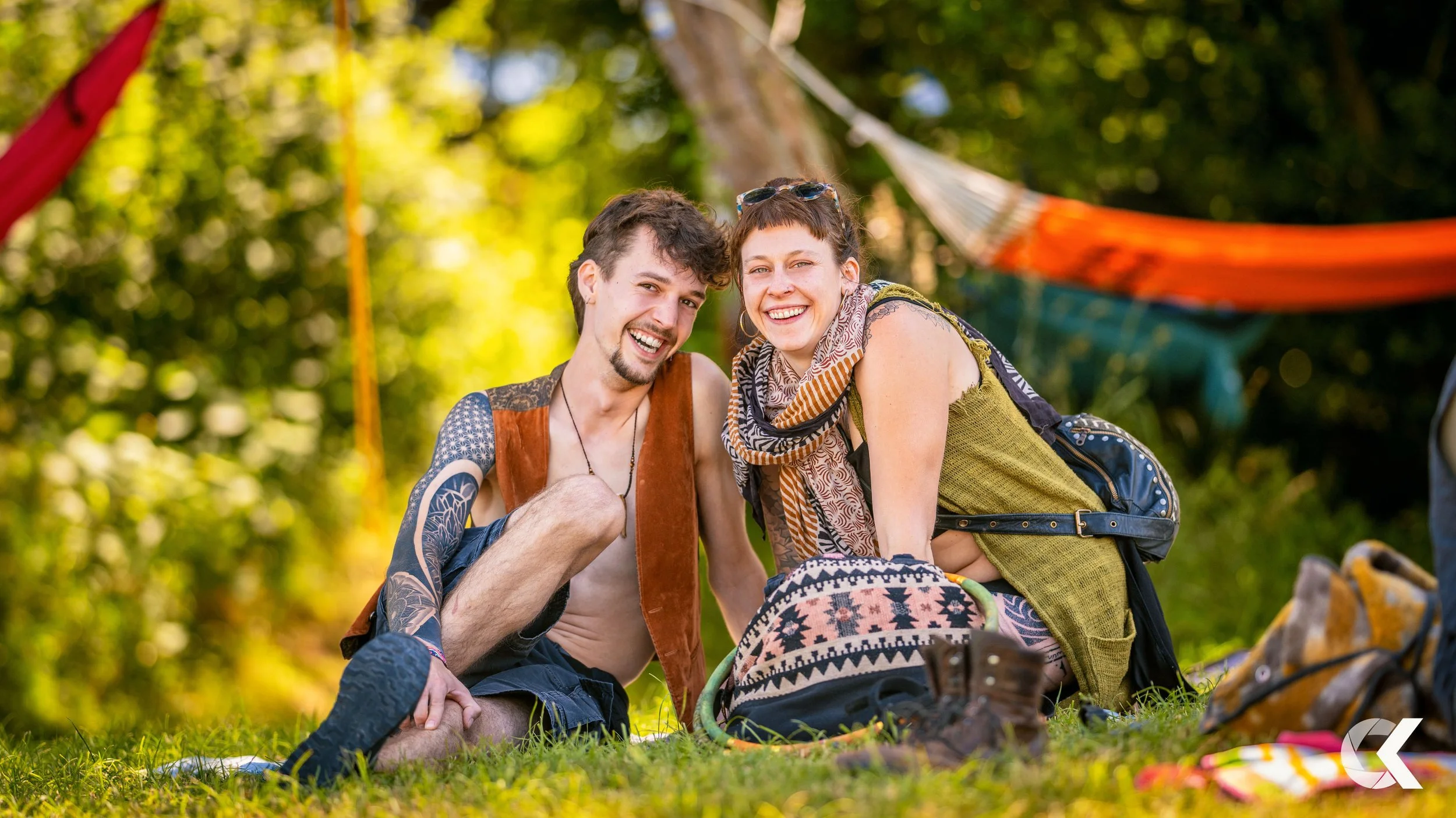 A smiling man and woman sitting on the grass in a wooded area with camping gear and hammocks in the background.