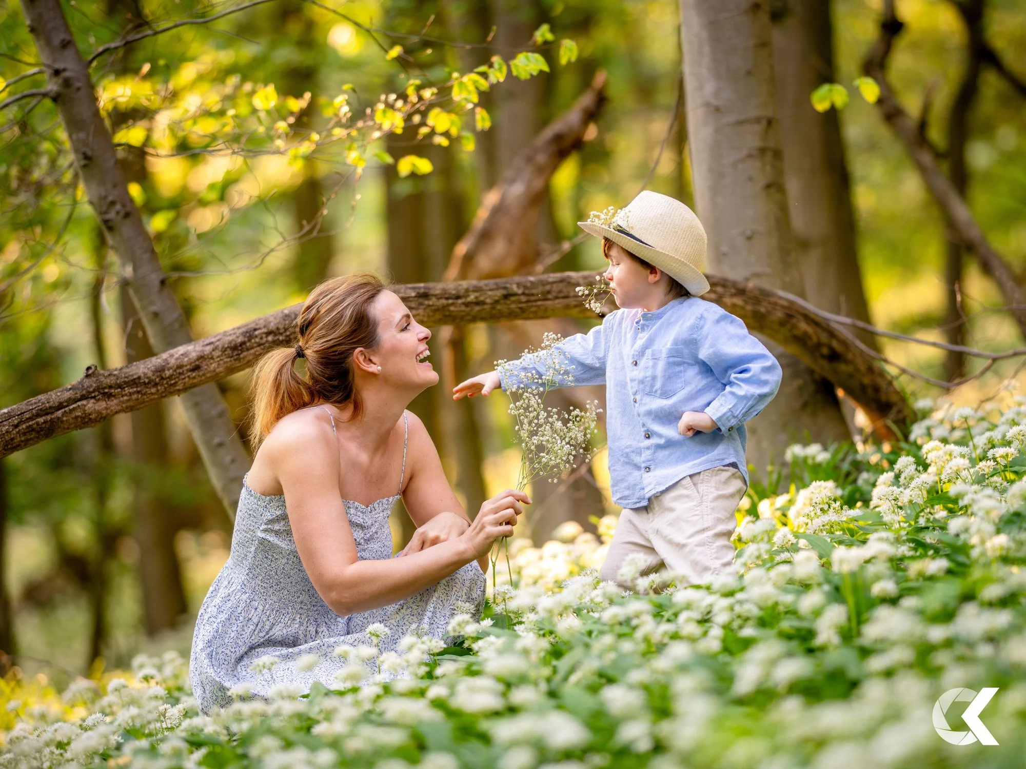 A woman and a young boy outdoors in a forested area filled with white flowers, the woman smiling and holding flowers while the boy, wearing a sunhat and blue shirt, reaches out to her.