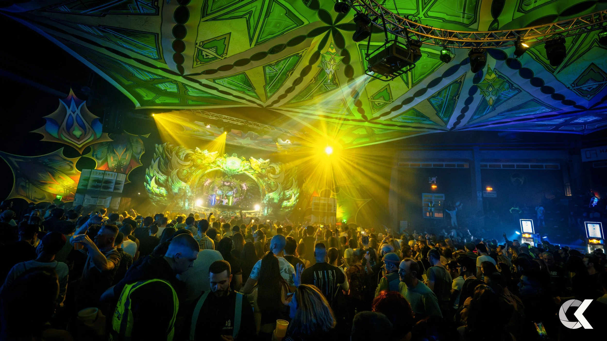 A crowded indoor concert with vibrant, colorful lighting and elaborate stage decorations, including a large, glowing lion face and intricate green and yellow patterns on the ceiling.