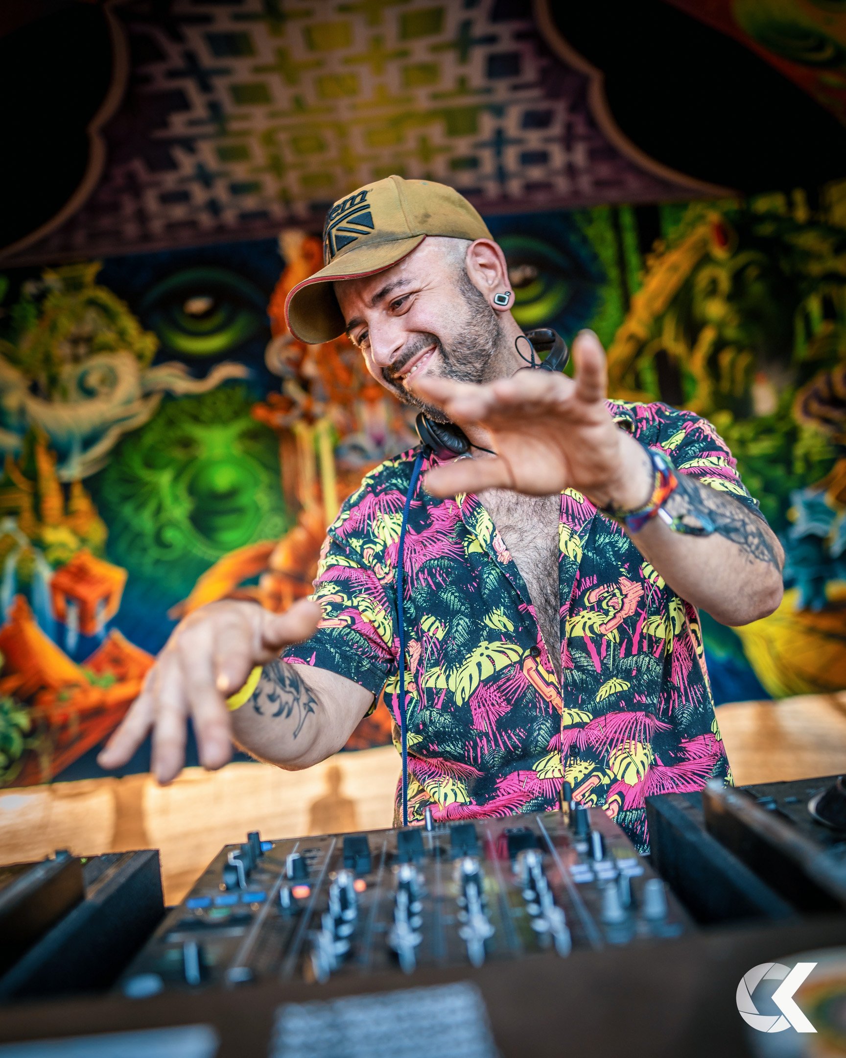 A DJ with a tropical shirt and a hat, smiling and gesturing with his hands while standing behind a DJ console, with colorful artistic mural in the background.