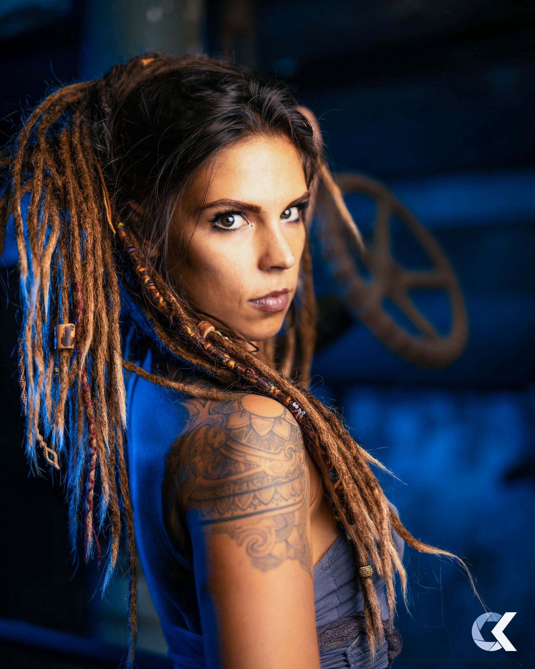 A woman with long dreadlocks and a large tattoo on her arm looking intently at the camera, with a dark background featuring metallic elements.