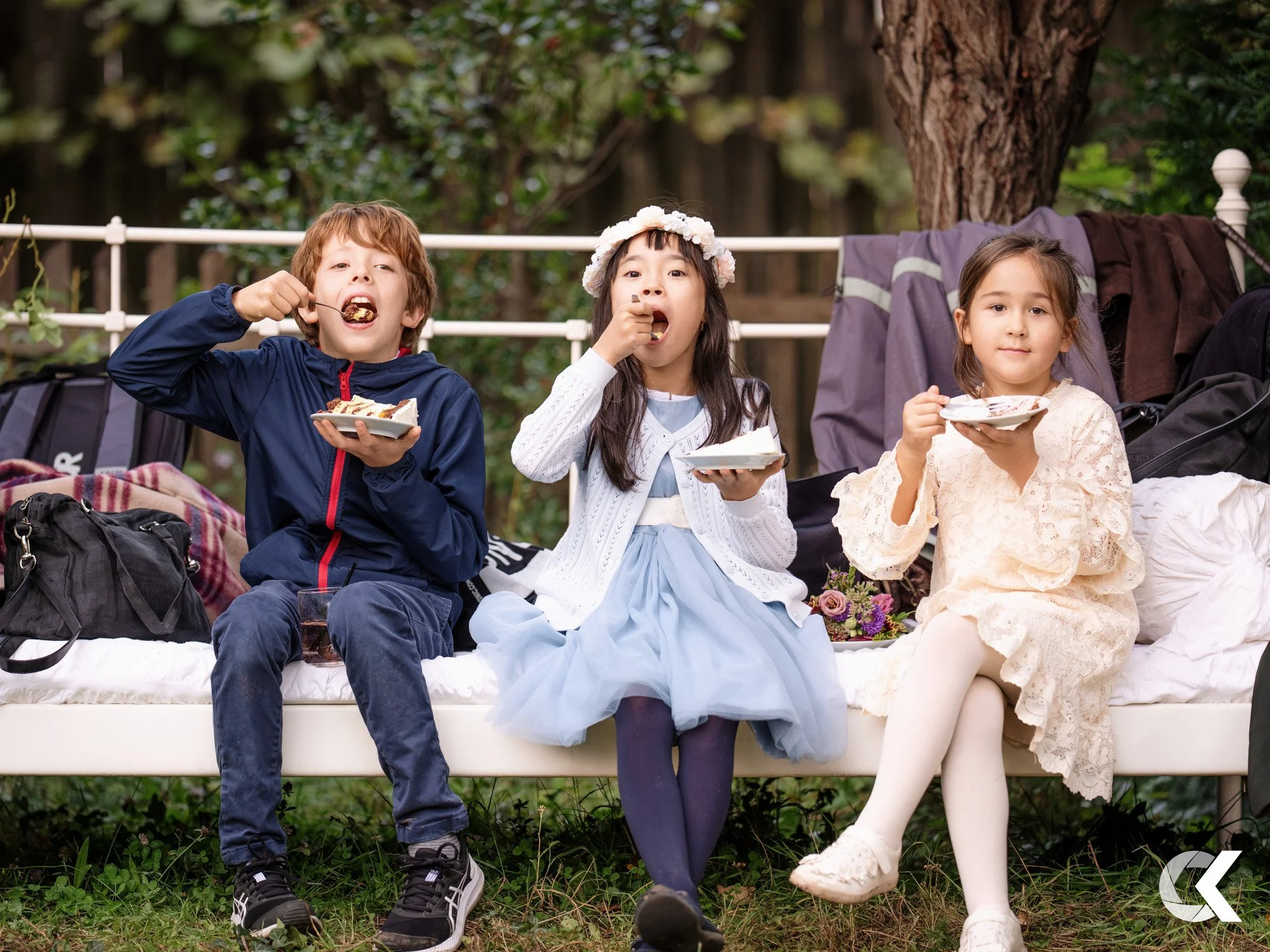 Three children sit on a white outdoor bench eating cake outdoors. The boy on the left wears a navy jacket and jeans, the girl in the middle wears a white cardigan and blue dress with a flower crown, and the girl on the right wears a cream lace dress.