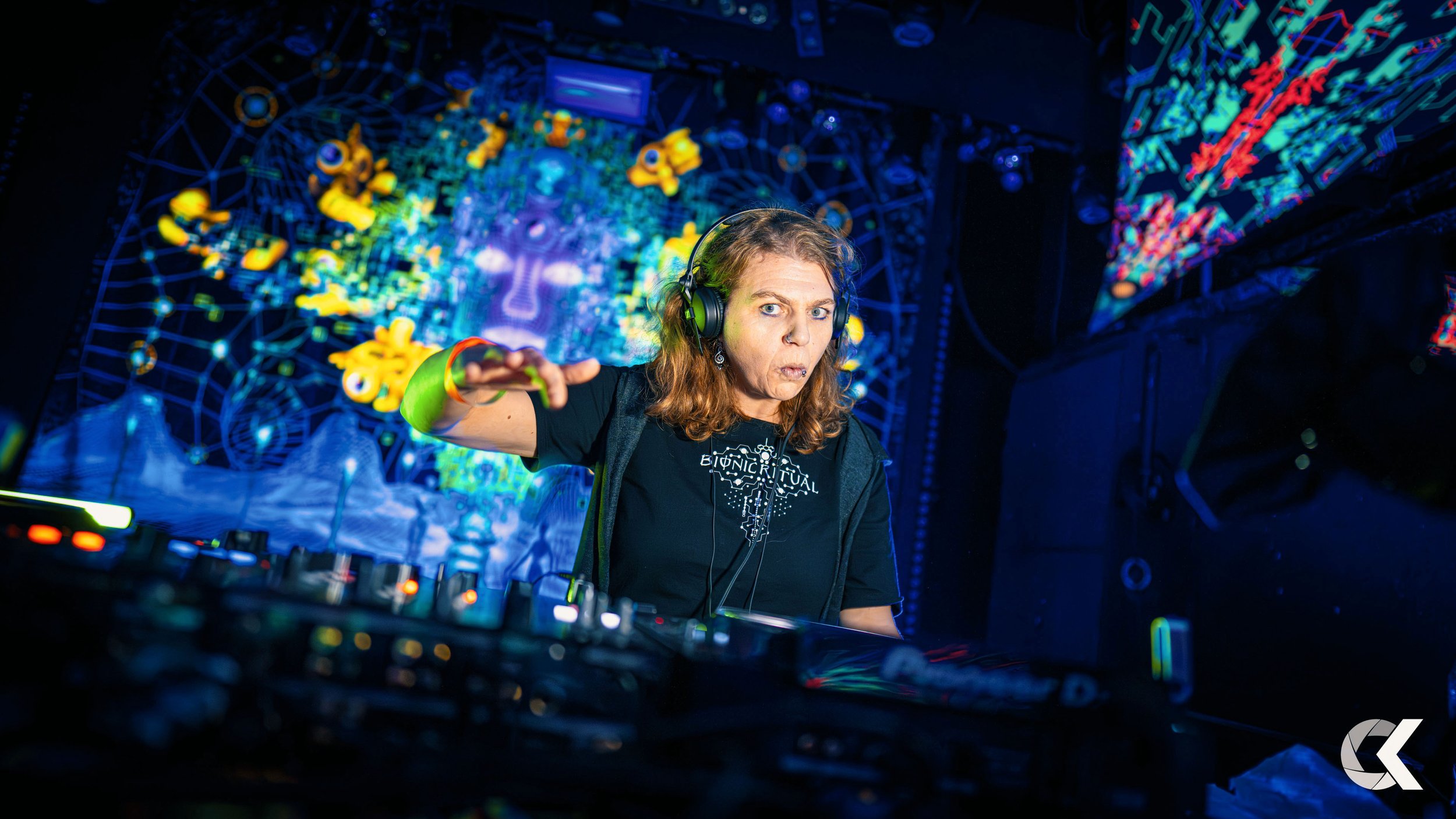 A female DJ with long hair, wearing headphones and a black shirt, is performing in a vibrant, colorful nightclub with digital art and geometric light displays in the background.