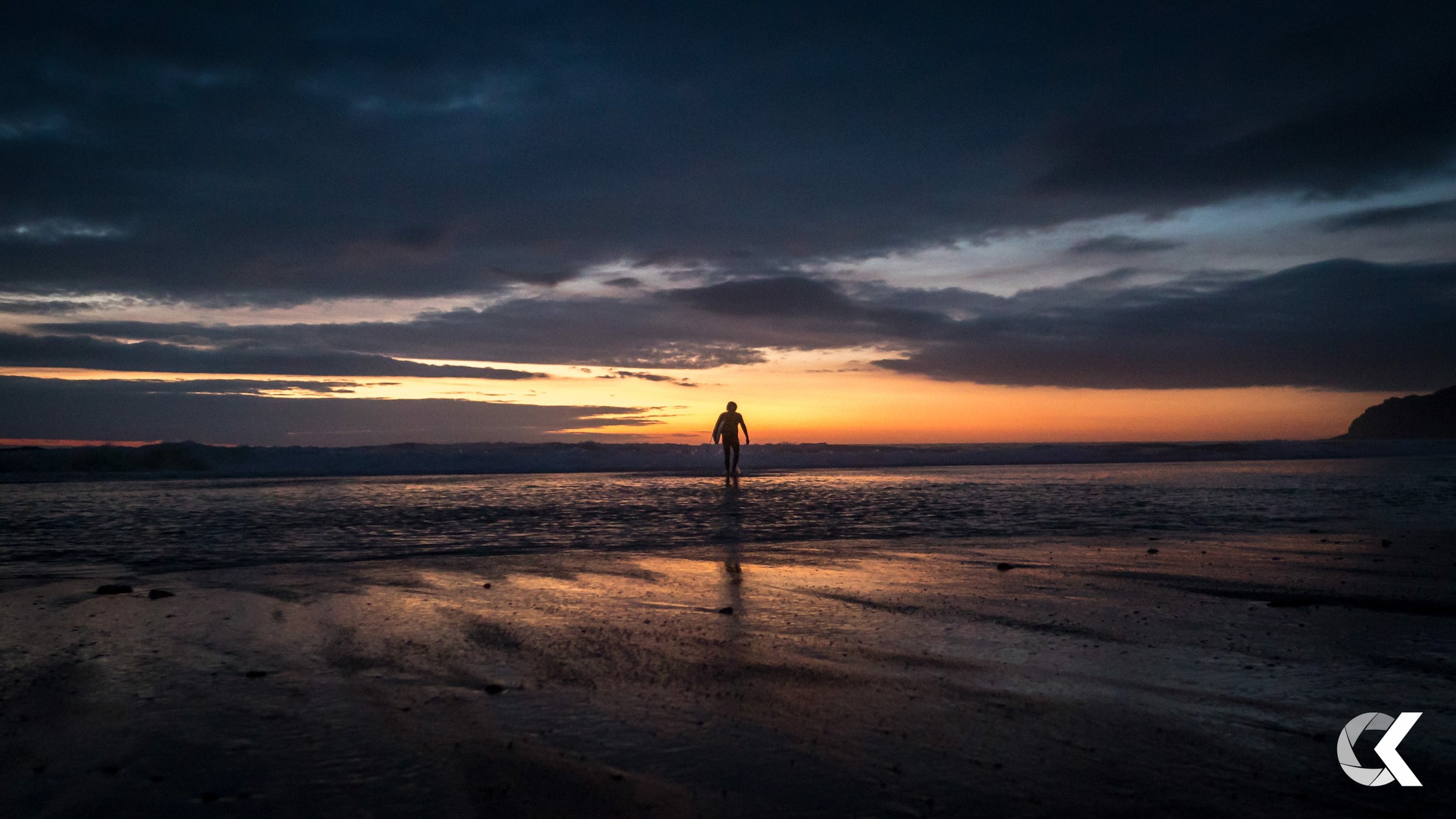 Person walking along the beach at sunset with the sky filled with dark clouds and orange hues, and reflections on wet sand.