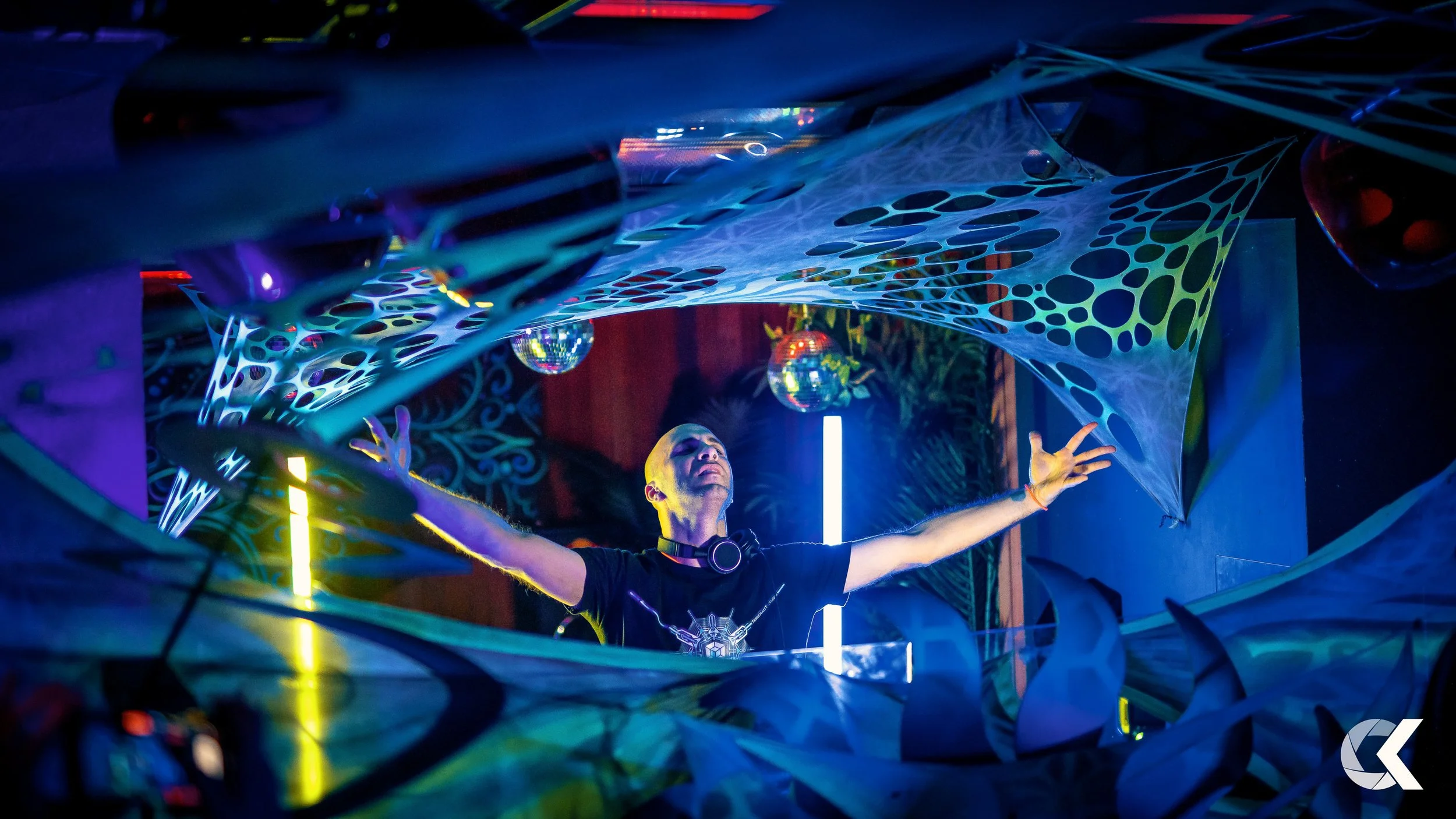 A DJ performing at a nightclub, with his arms outstretched and eyes closed. The scene includes colorful lighting, hanging disco balls, and futuristic decorations with web-like structures.