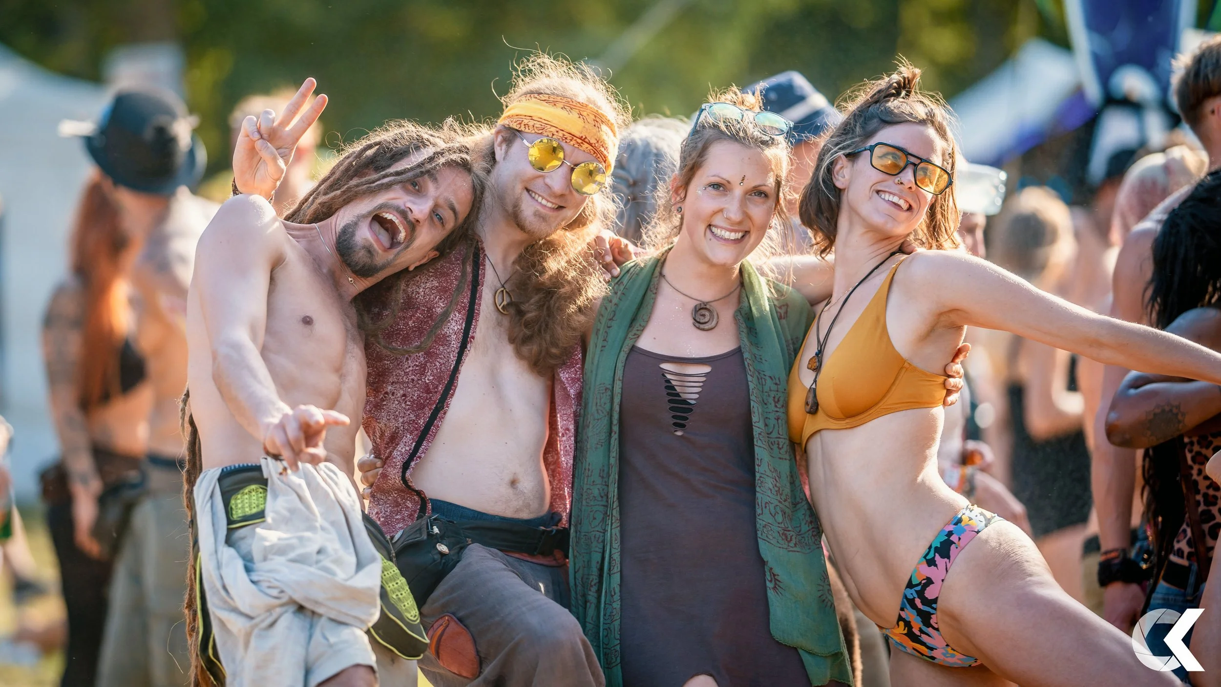 Four friends at a music festival, smiling and embracing each other, with people and tents in the background.