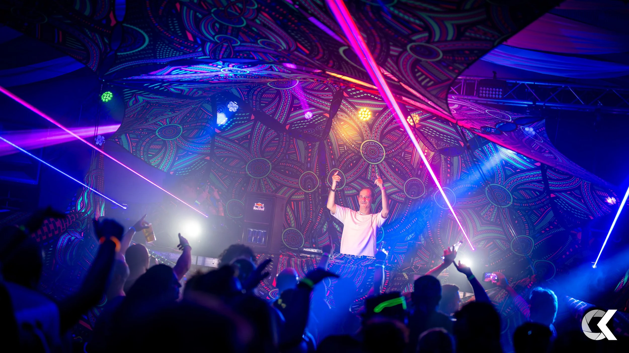 A DJ is performing on a stage with colorful psychedelic backdrop and vibrant laser lights, crowd dancing and enjoying the music inside a lively nightclub.
