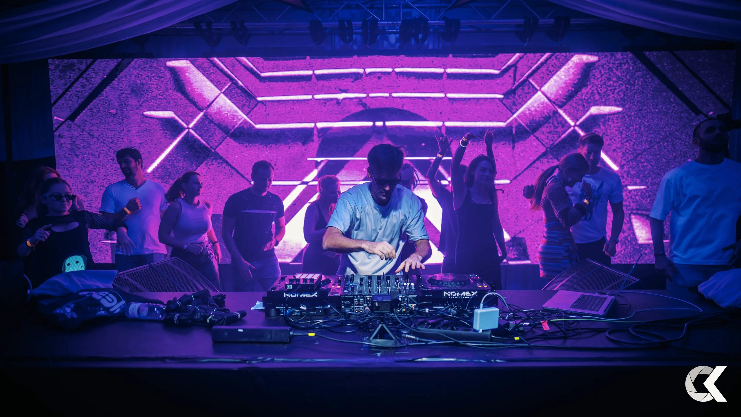 A DJ performing on stage in front of a large purple LED screen with people dancing and enjoying the music.