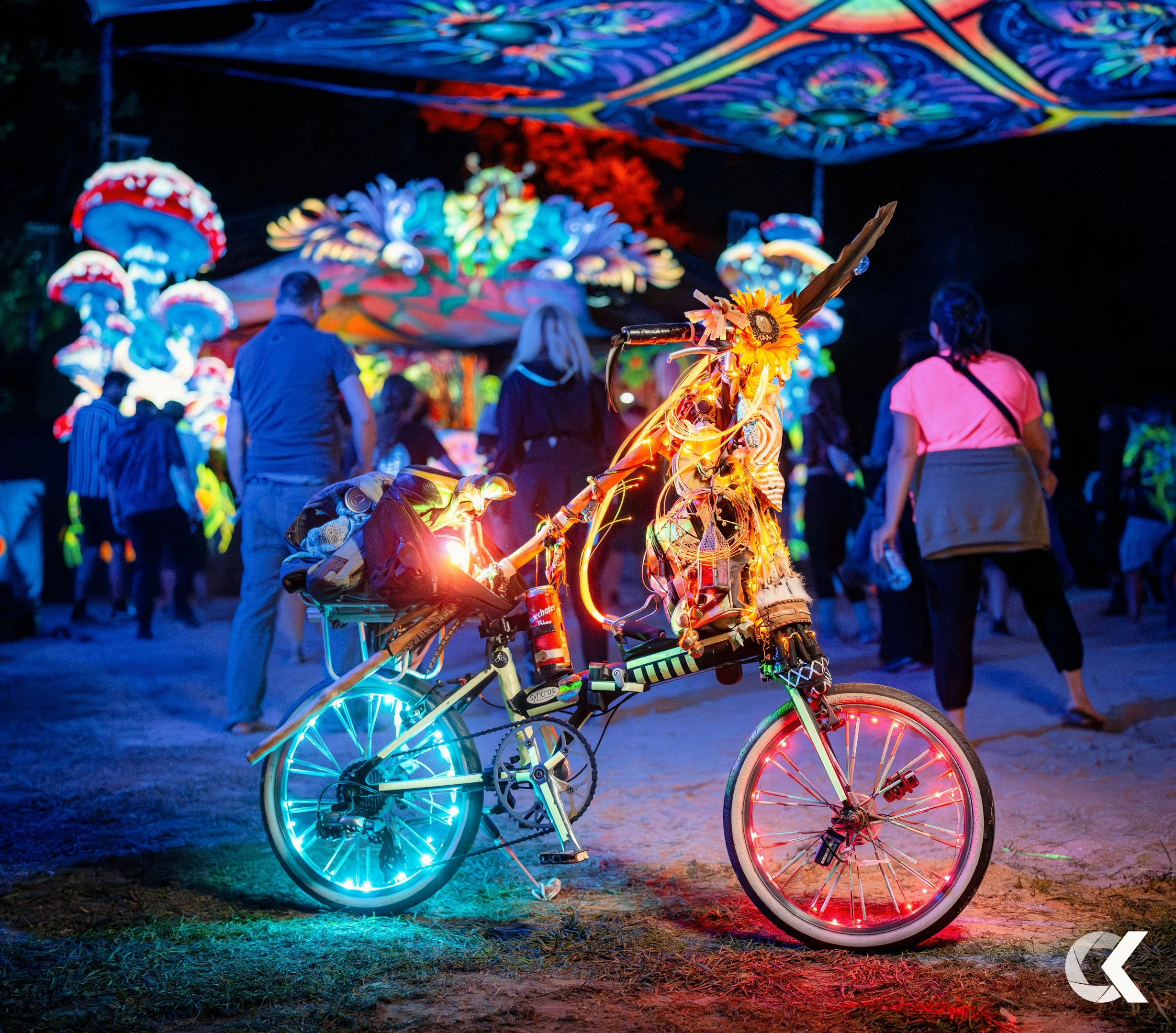 Decorated bicycle with multicolored lights and accessories at a nighttime festival, with people walking and illuminated art installations in the background.