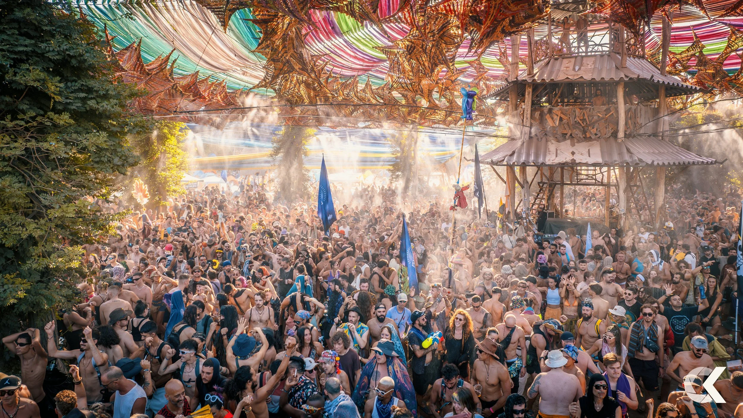 A crowded outdoor festival with many people dancing and enjoying music, colorful decorations overhead, and a wooden stage structure in the background.