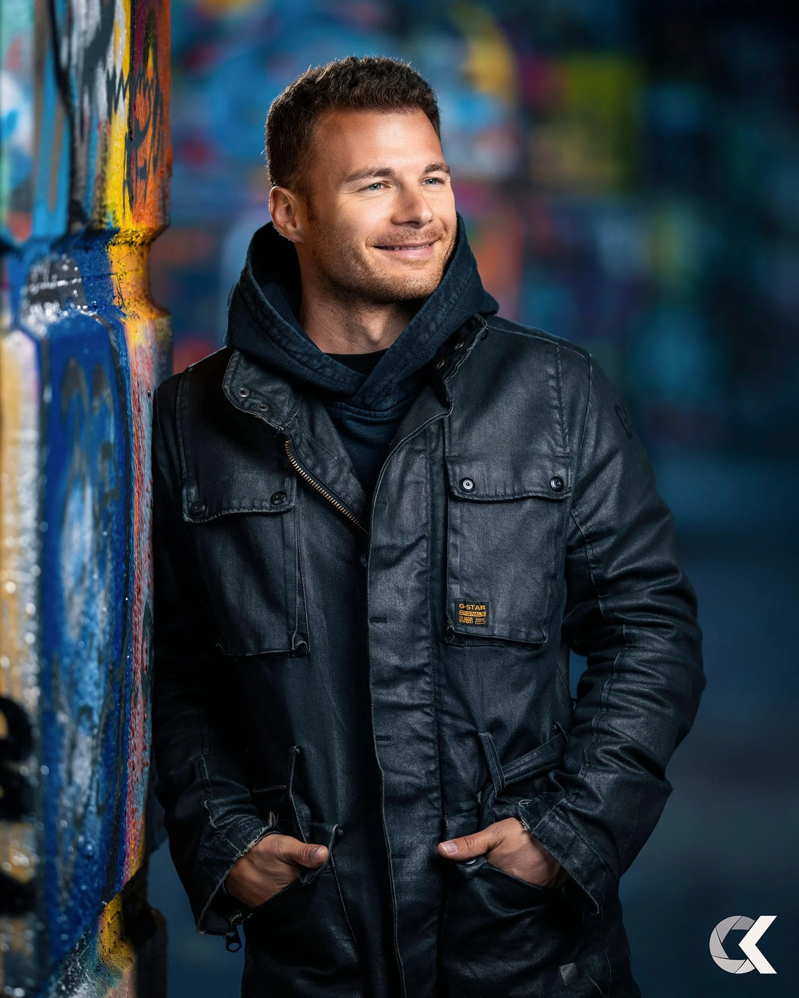 Ein smiling Mann mit kurzen braunen Haaren steht neben einer bunten Graffiti-Wand, trägt eine schwarze Lederjacke und eine schwarze Kapuzenjacke darunter, mit den Händen in den Taschen.