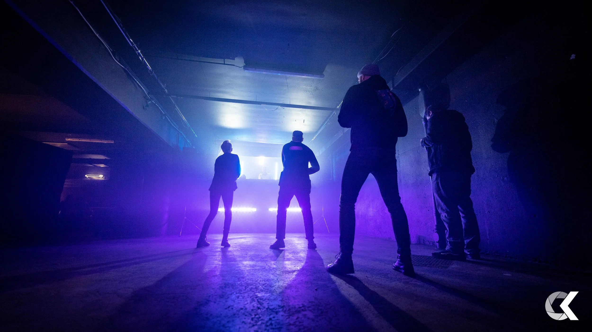 Four people standing in a dark, underground location with purple and blue lighting, facing a DJ on a raised platform.
