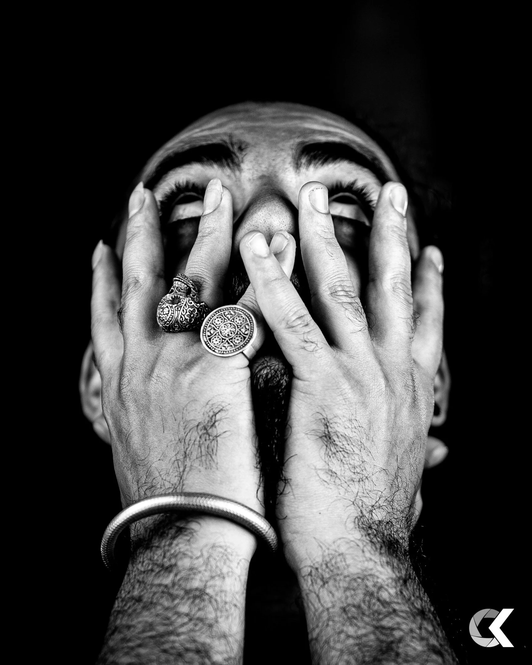 A black and white photo of a person's face partially covered by their hands, showing expressive facial features, thick eyebrows, and rings on fingers, with a dark background.