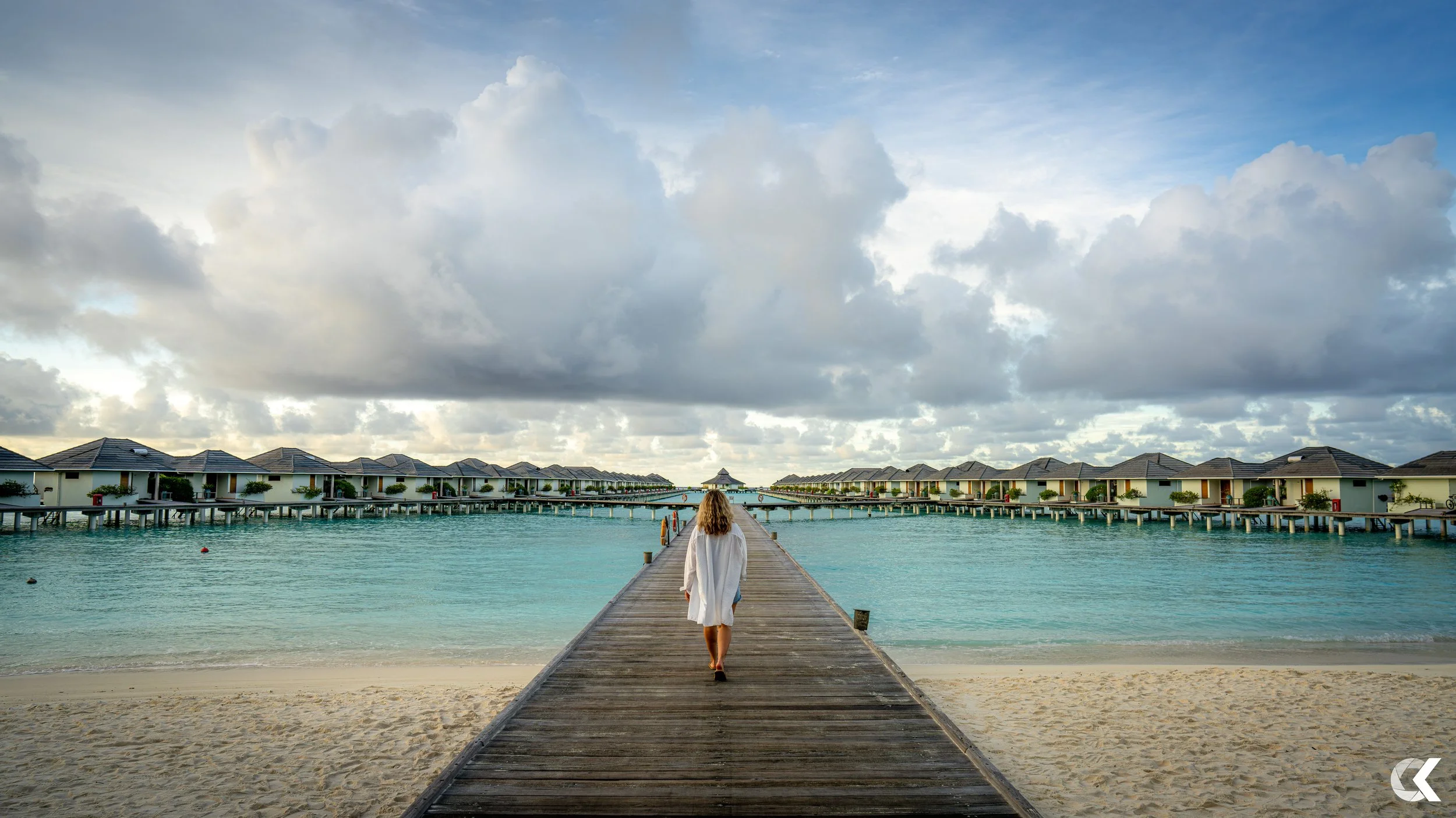 A woman walking on a wooden pier towards overwater bungalows on a tropical beach with cloudy sky.