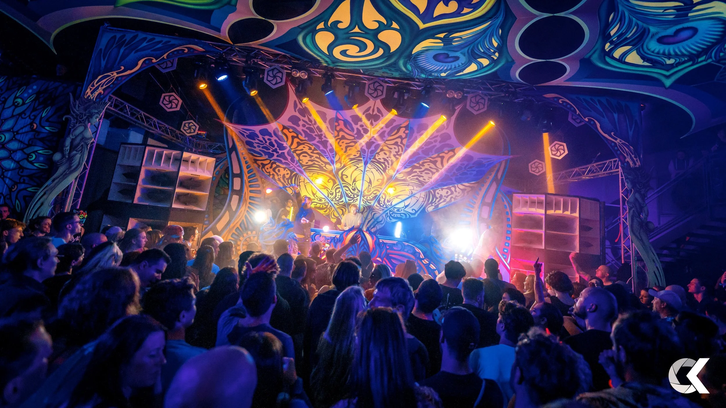 People dancing at a colorful indoor concert or music festival with vibrant lights and graffiti-style artwork on the ceiling and stage backdrop.