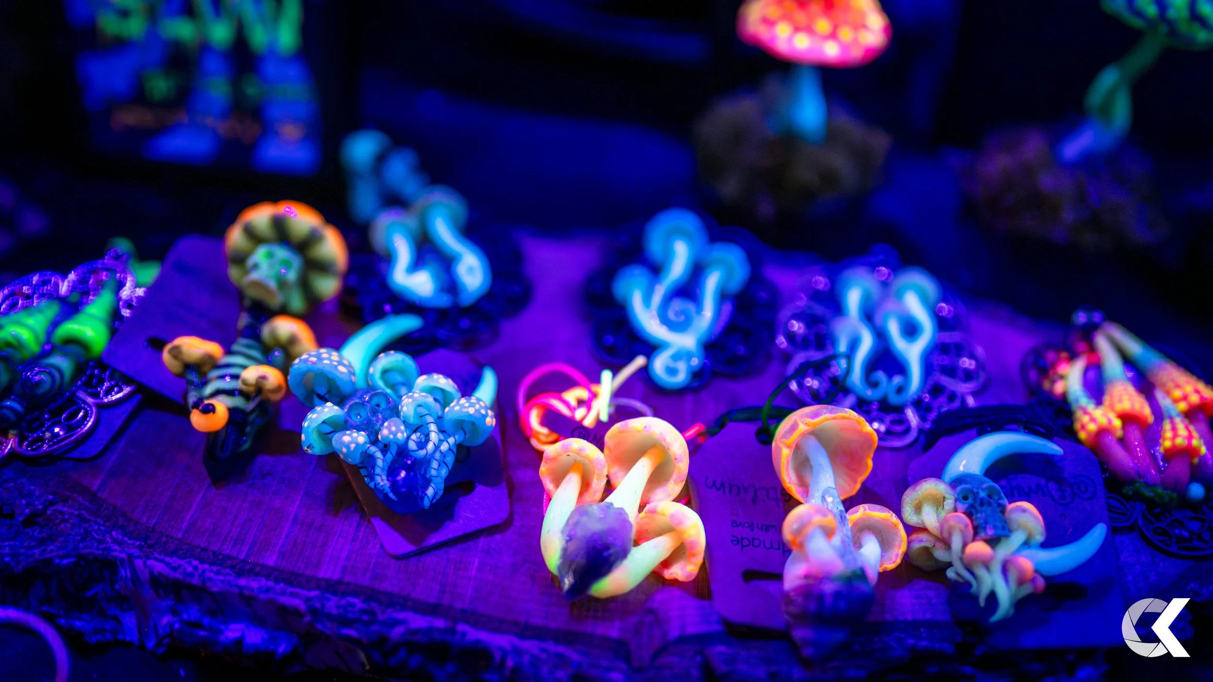Colorful glow-in-the-dark jewelry and accessories on display on a wooden surface under blacklight, including rings, earrings, and necklaces with neon colors and mushroom-shaped designs.