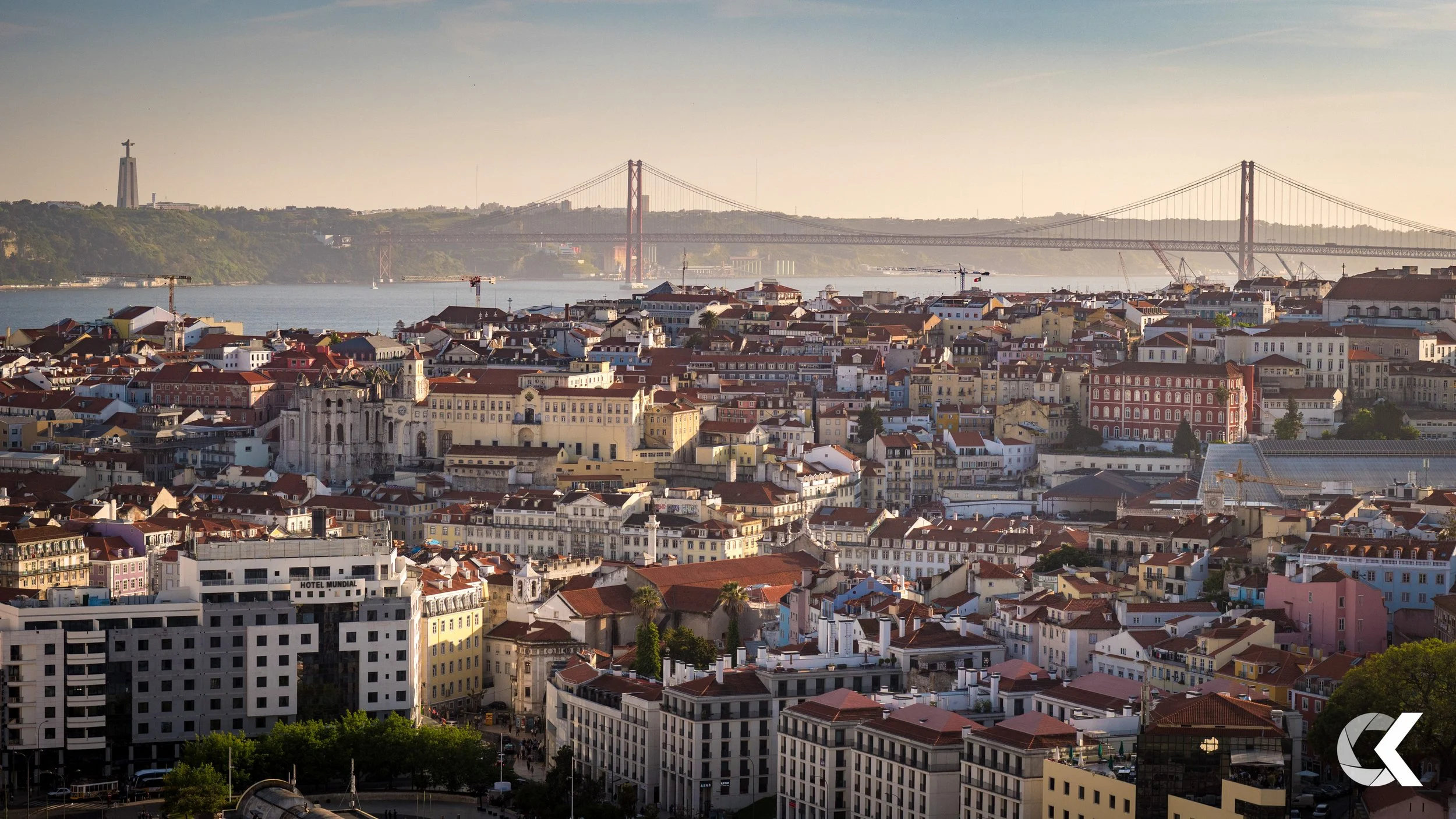 A cityscape featuring a densely packed urban area with various buildings, a river in the background, and a large suspension bridge crossing the river. The scene is taken from an elevated viewpoint during daylight.