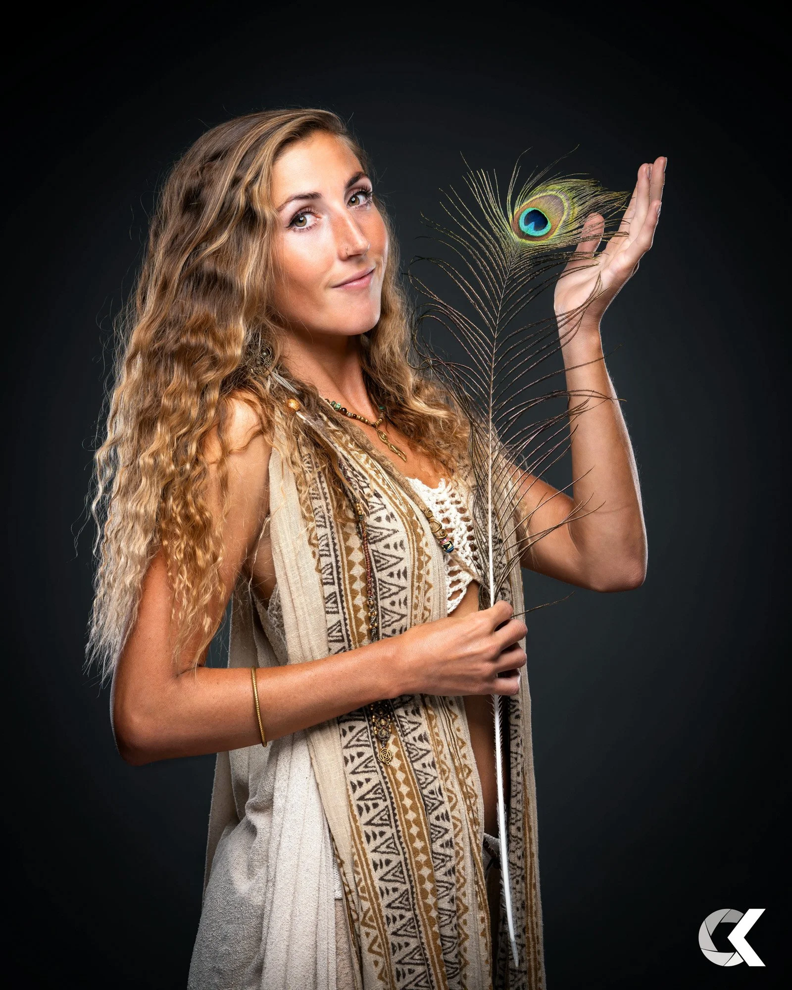 A woman with long, curly blond hair holding a peacock feather against a dark background.