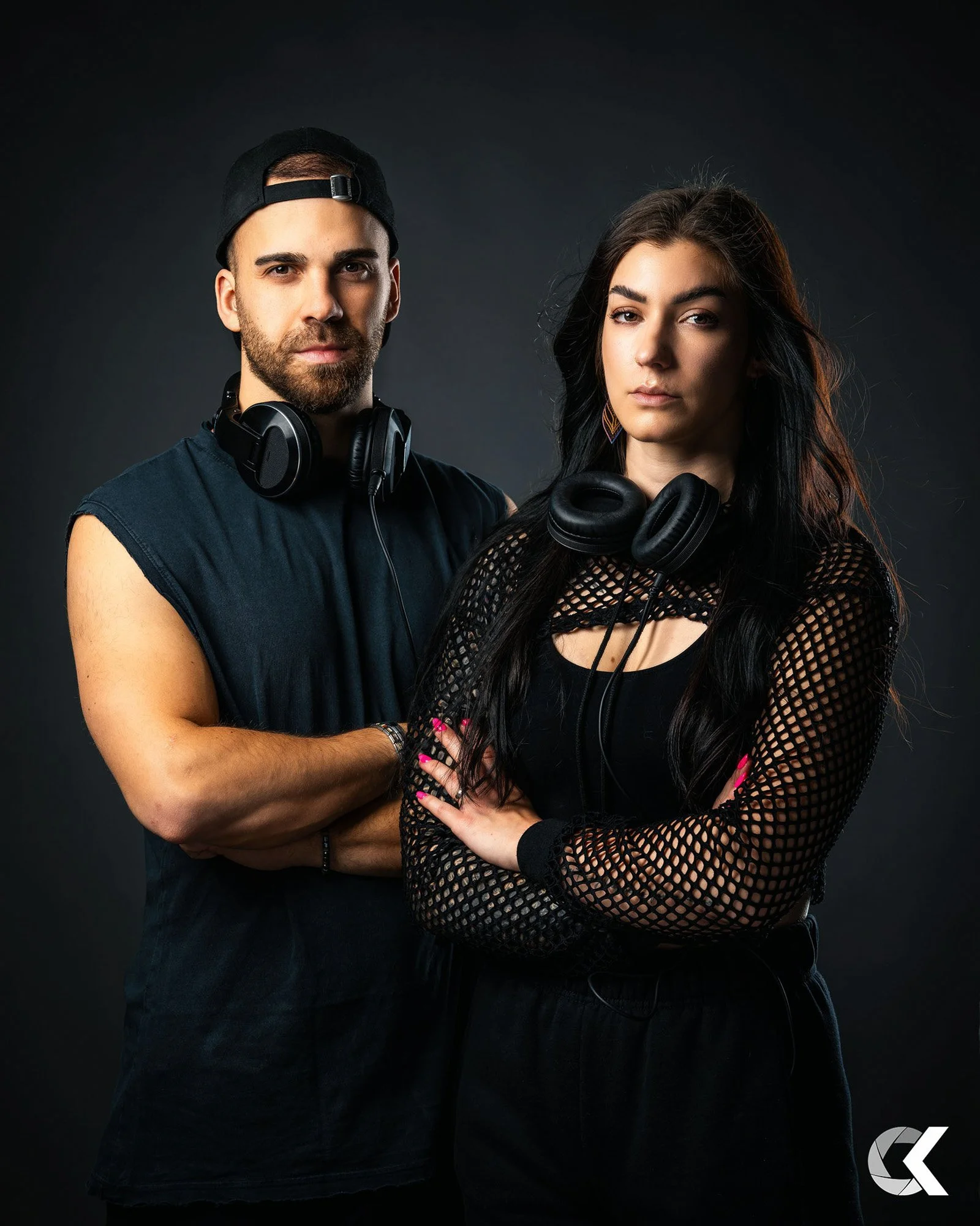 A young man and woman standing with arms crossed, wearing black clothing and headphones, against a dark background.