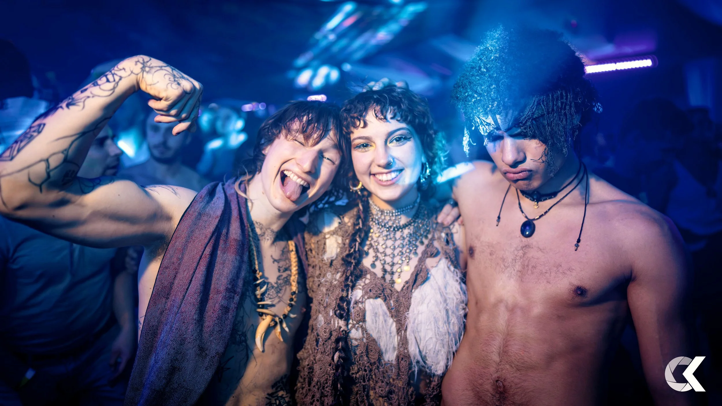 Three young people at a party, standing close together and smiling. One person is flexing their arm, another is sticking out their tongue, and the third is looking down. The scene is vibrant with blue and purple lighting.