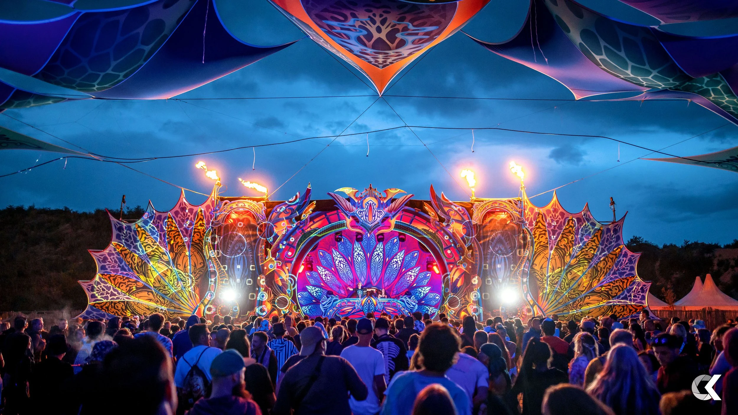 A large crowd at an outdoor music festival during evening with a colorful, illuminated stage featuring artistic, butterfly-inspired designs, fire effects, and a DJ performing, under a darkening sky.