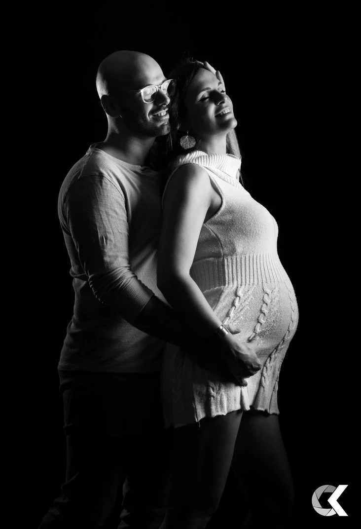 A black and white photo of a pregnant woman and a man standing behind her, smiling and embracing, with her hand resting on her baby bump.
