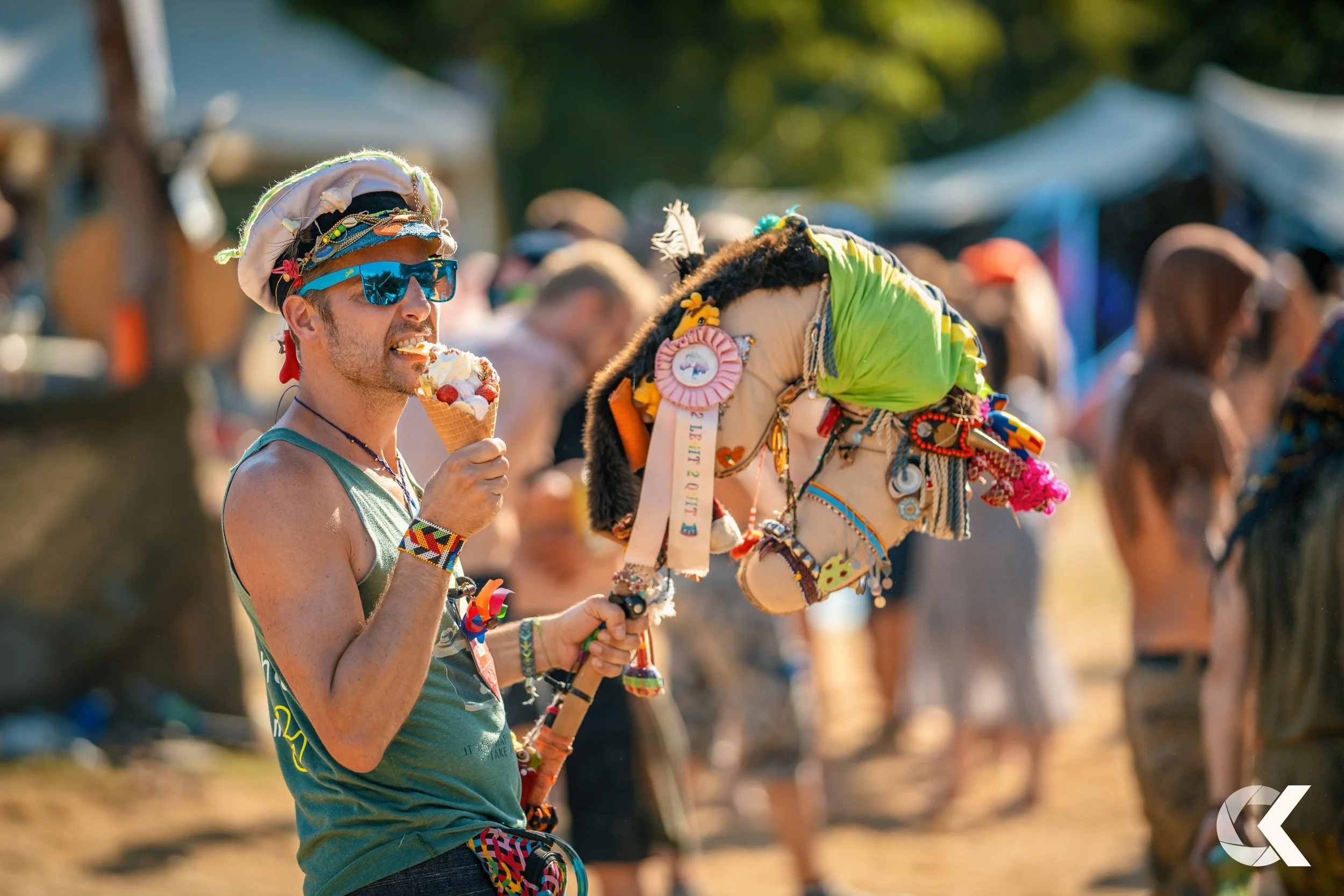 A person wearing sunglasses, a hand-made hat, and colorful jewelry, holding a decorated stick and eating ice cream at an outdoor festival or gathering.