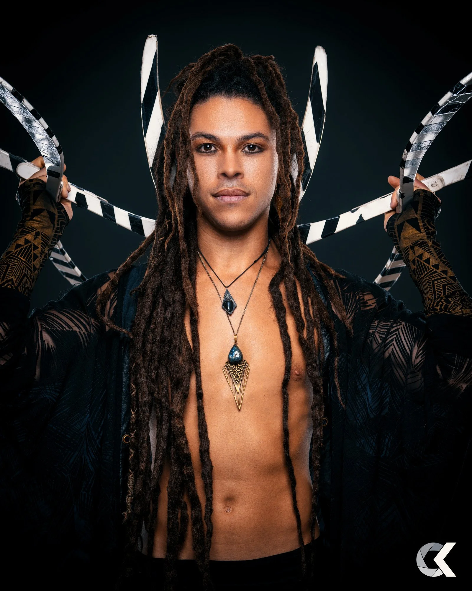A person with long dreadlocks wearing a black lace top, holding a black and white striped prop behind their head, against a dark background, with necklaces featuring blue stones.