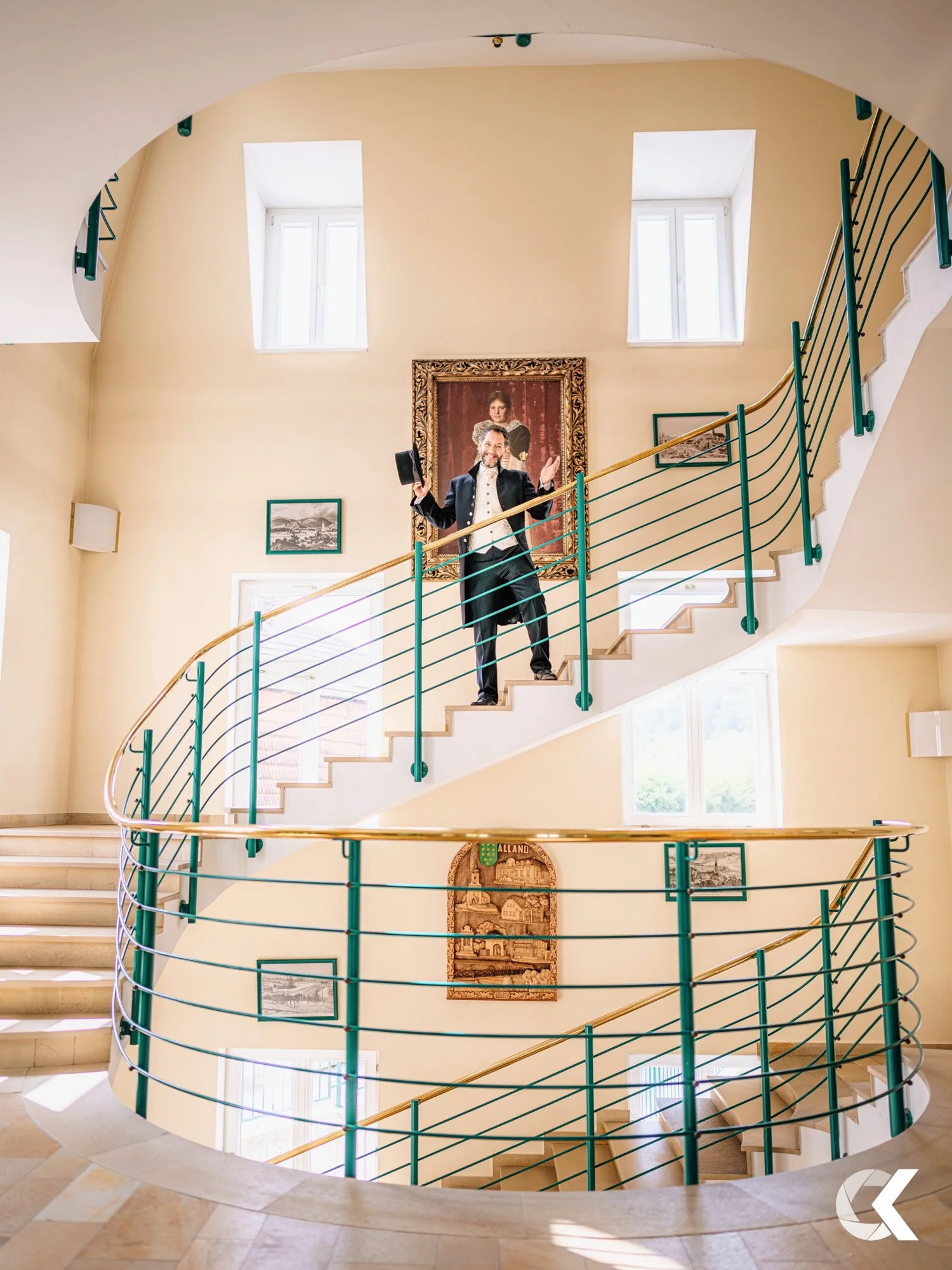 A man dressed in a tuxedo standing on a spiral staircase in a well-lit interior, holding a top hat and waving. The staircase has green railings and wooden handrails, with framed photographs and artwork on the cream-colored walls behind him.