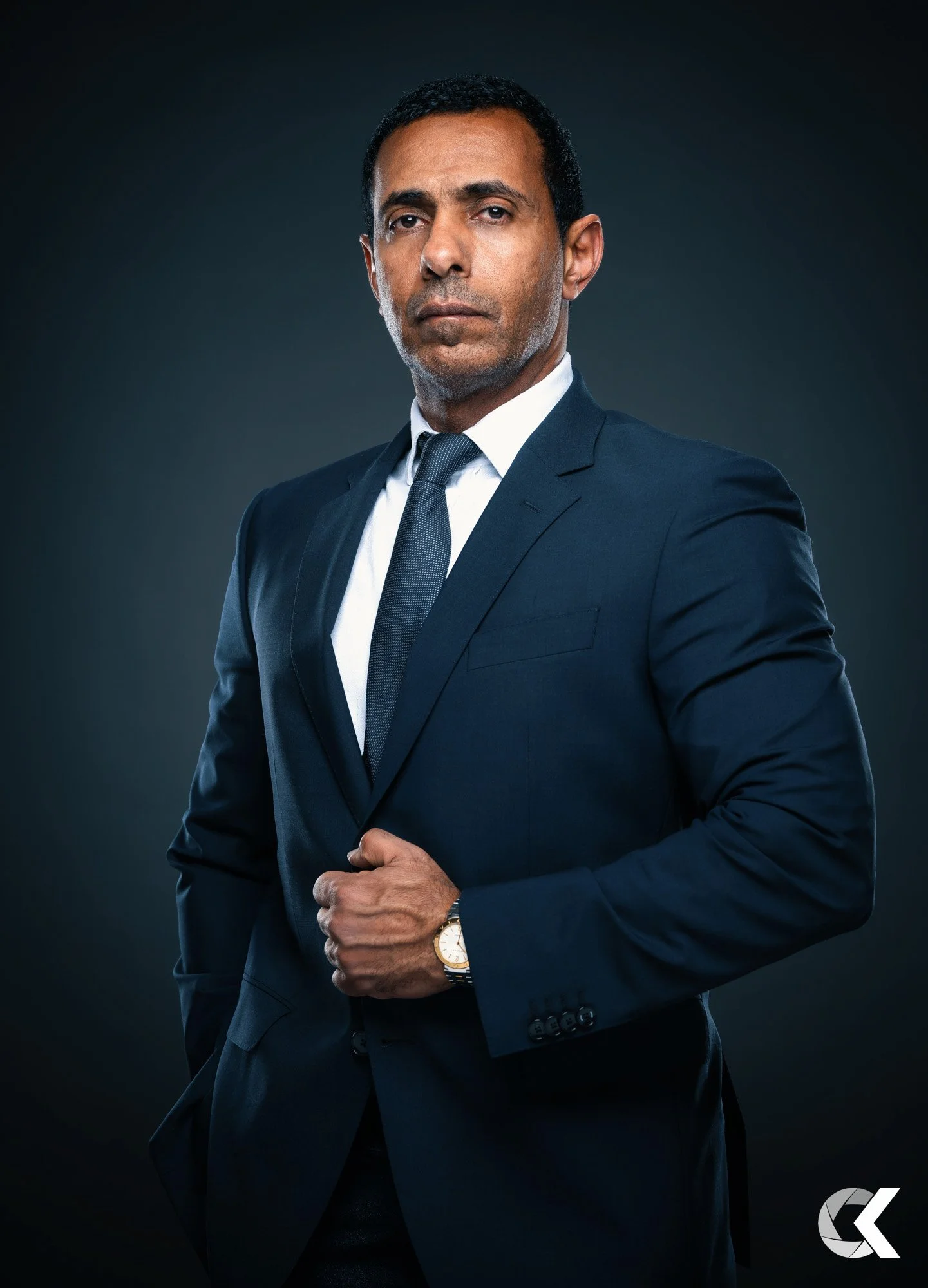 A man in a dark blue suit, white shirt, and dark tie standing against a dark background. He has short black hair, medium skin, and is looking directly at the camera with a serious expression. His left hand is adjusting his suit jacket, and he is wear