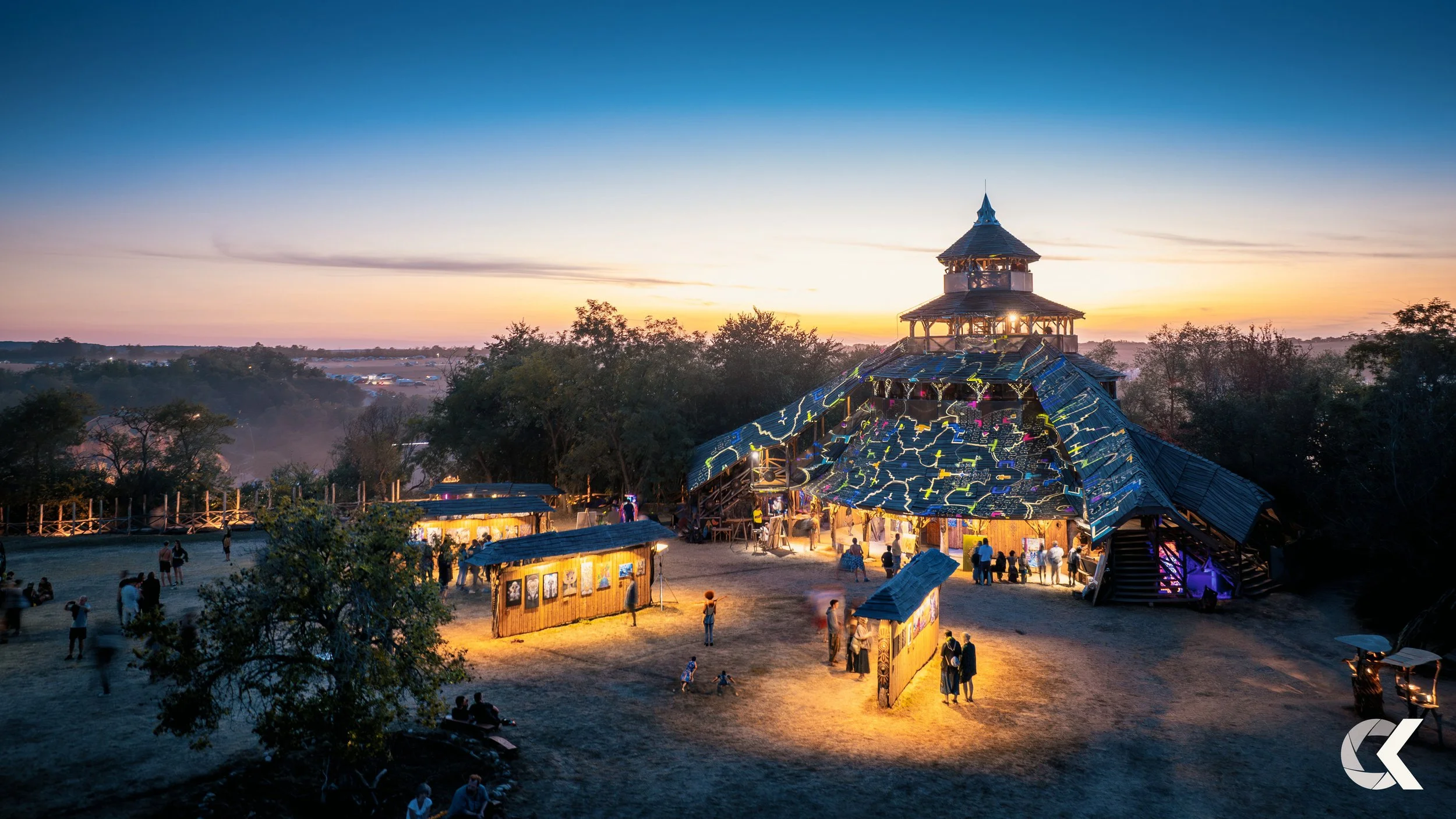An outdoor event at sunset with a large wooden structure decorated with colorful lights, surrounded by smaller booths and people walking and socializing on a sandy ground.