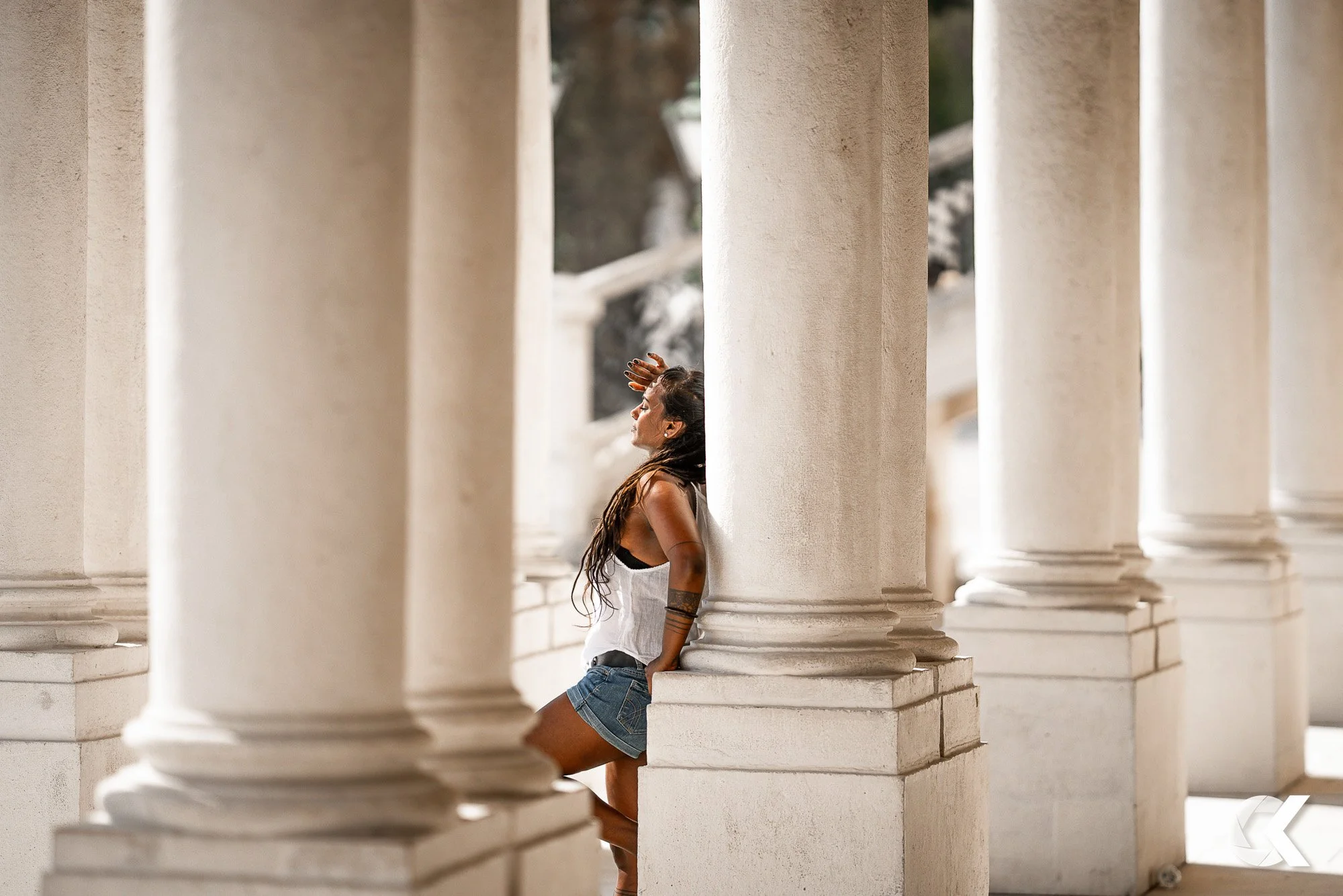 A woman leaning against a large column in a classical architectural setting, with multiple columns surrounding her and sunlight shining on her face.