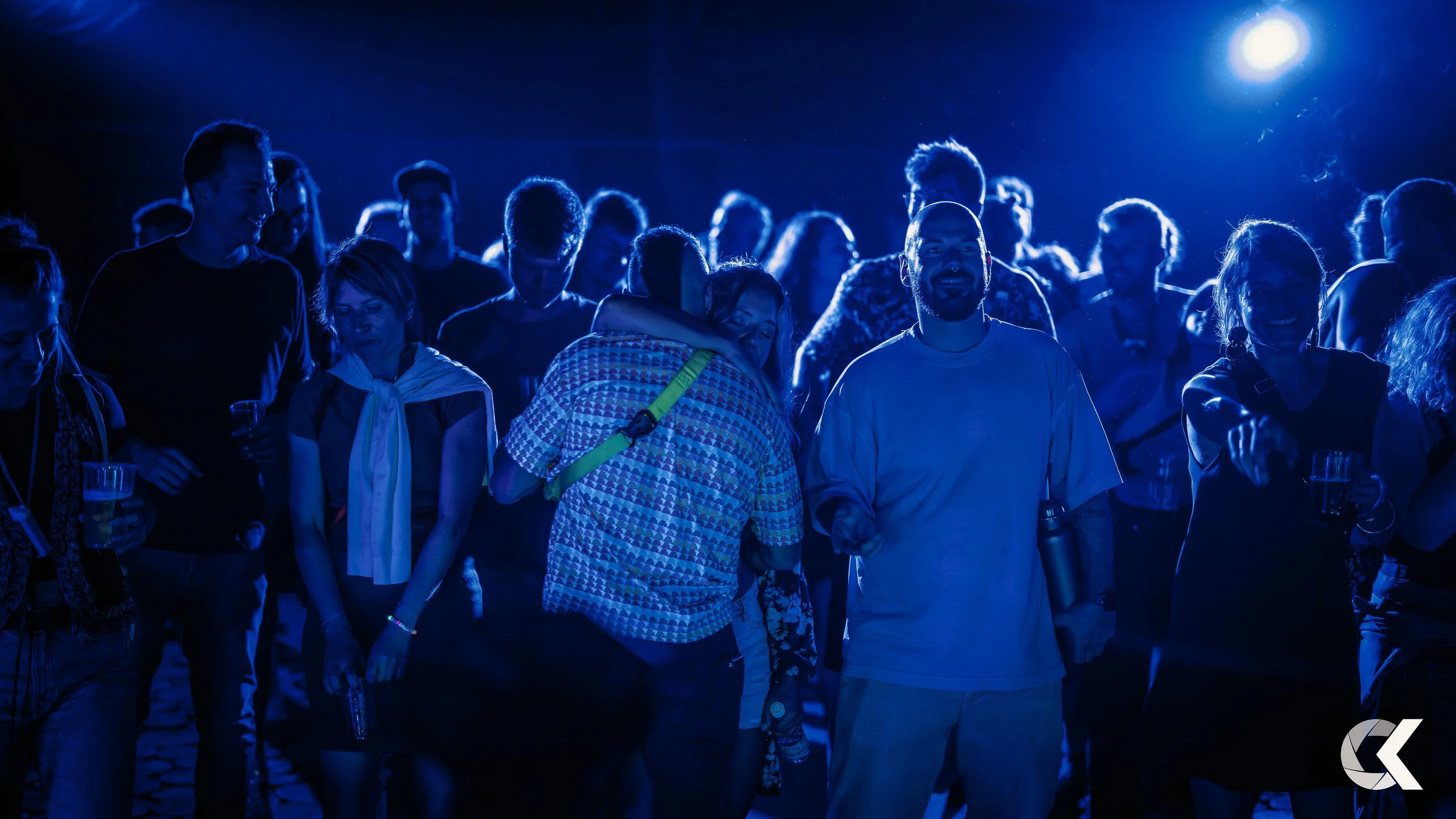 A group of people at a nighttime outdoor concert or event, illuminated by blue lighting, some smiling and others engaging with each other, holding drinks.