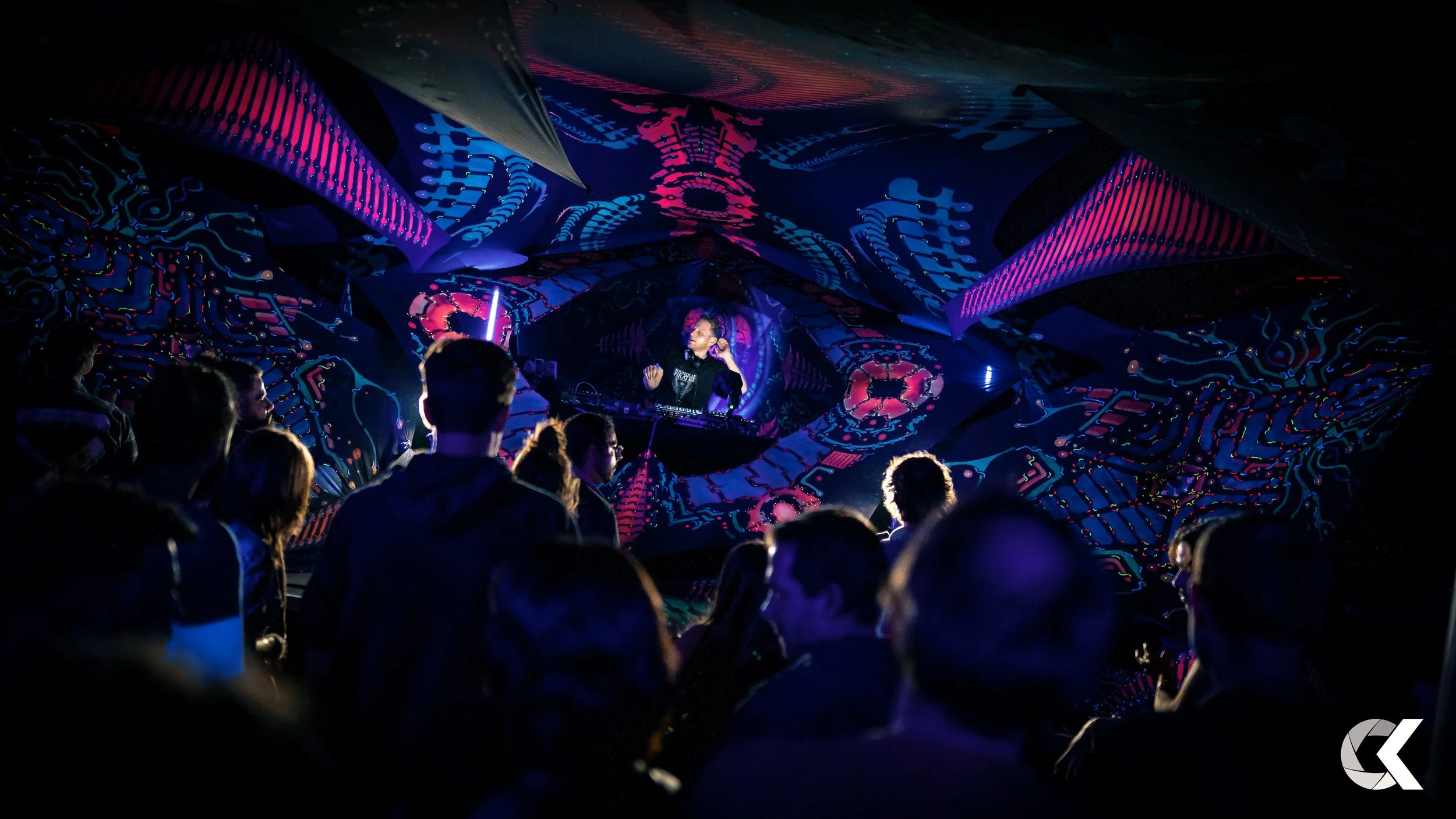 A DJ performs at a digital art event with neon and LED light projections on the ceiling and walls. A crowd of people watches the performance in a dark room illuminated by colorful light displays.