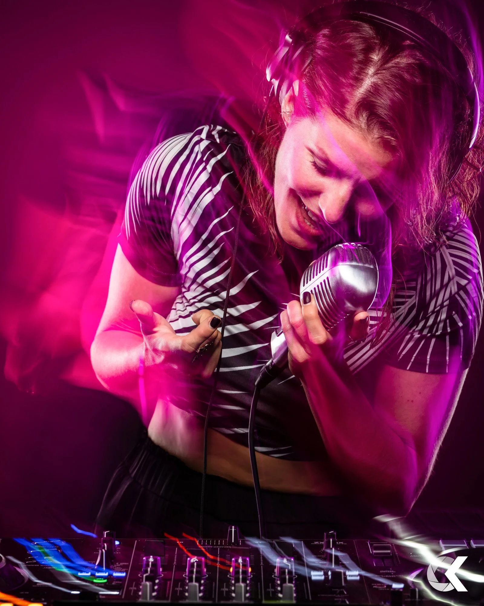 Junge Frau mit Kopfhörern beim Singen oder Musikhören, im Hintergrund DJ-Equipment, mithilfe von bunt leuchtenden Lichtern, violet und pink, bei einem Live-Auftritt oder Party.