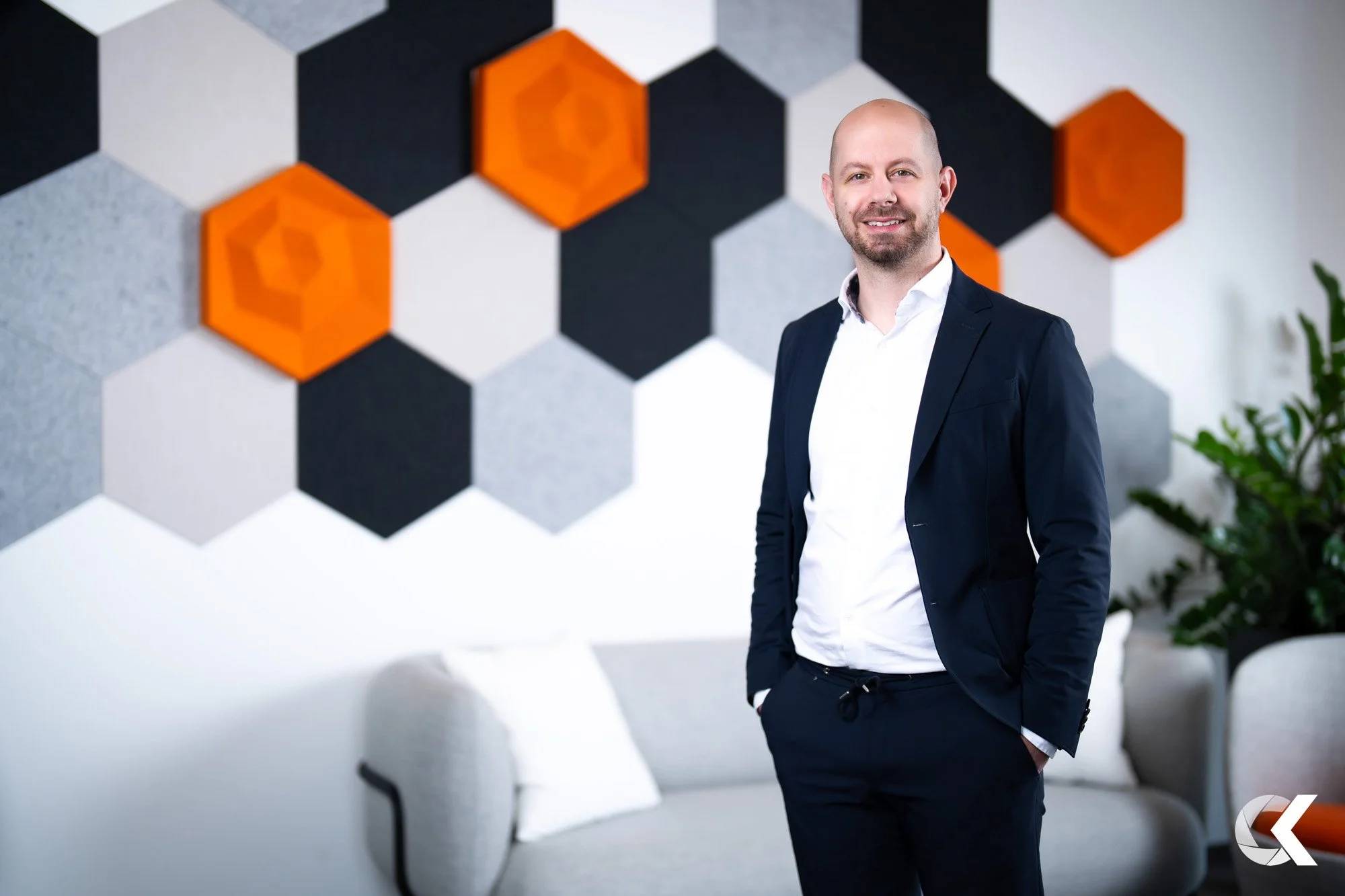 A man in a navy blue suit standing in a modern office or lounge with a colorful hexagonal wall art and a beige sofa with pillows. There's a green plant on the right.